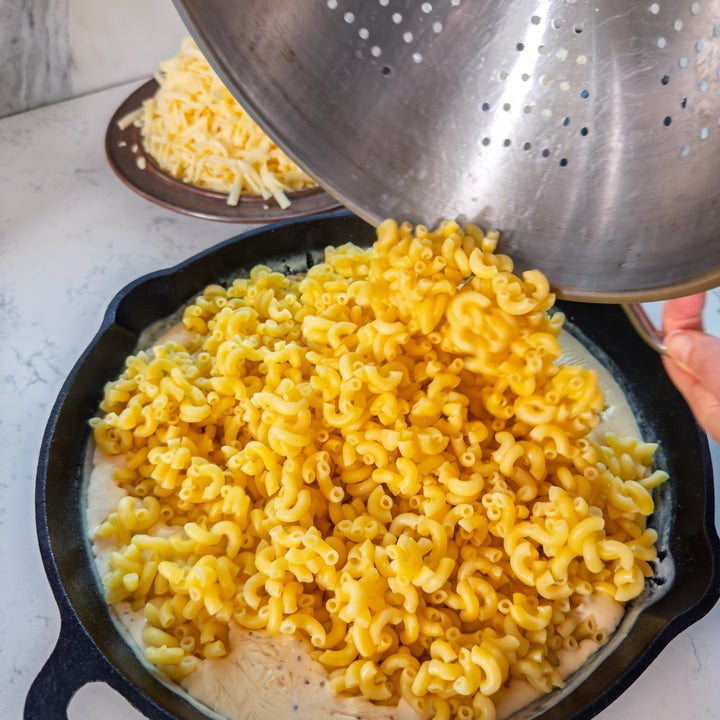 Cooked pasta is being poured from a colander into a skillet with cheese sauce. A plate of grated cheese is in the background