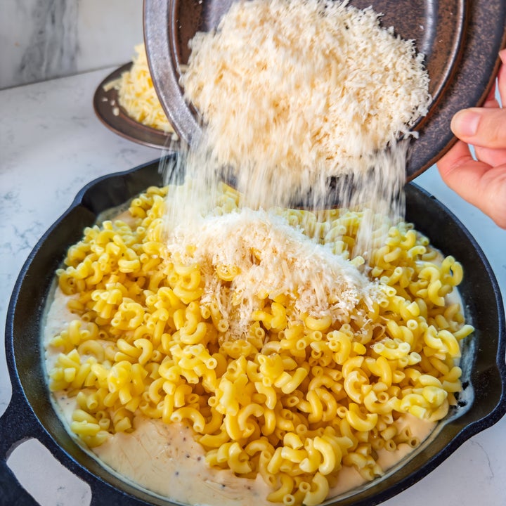 Cheese is being added to a skillet filled with cooked elbow macaroni, preparing macaroni and cheese