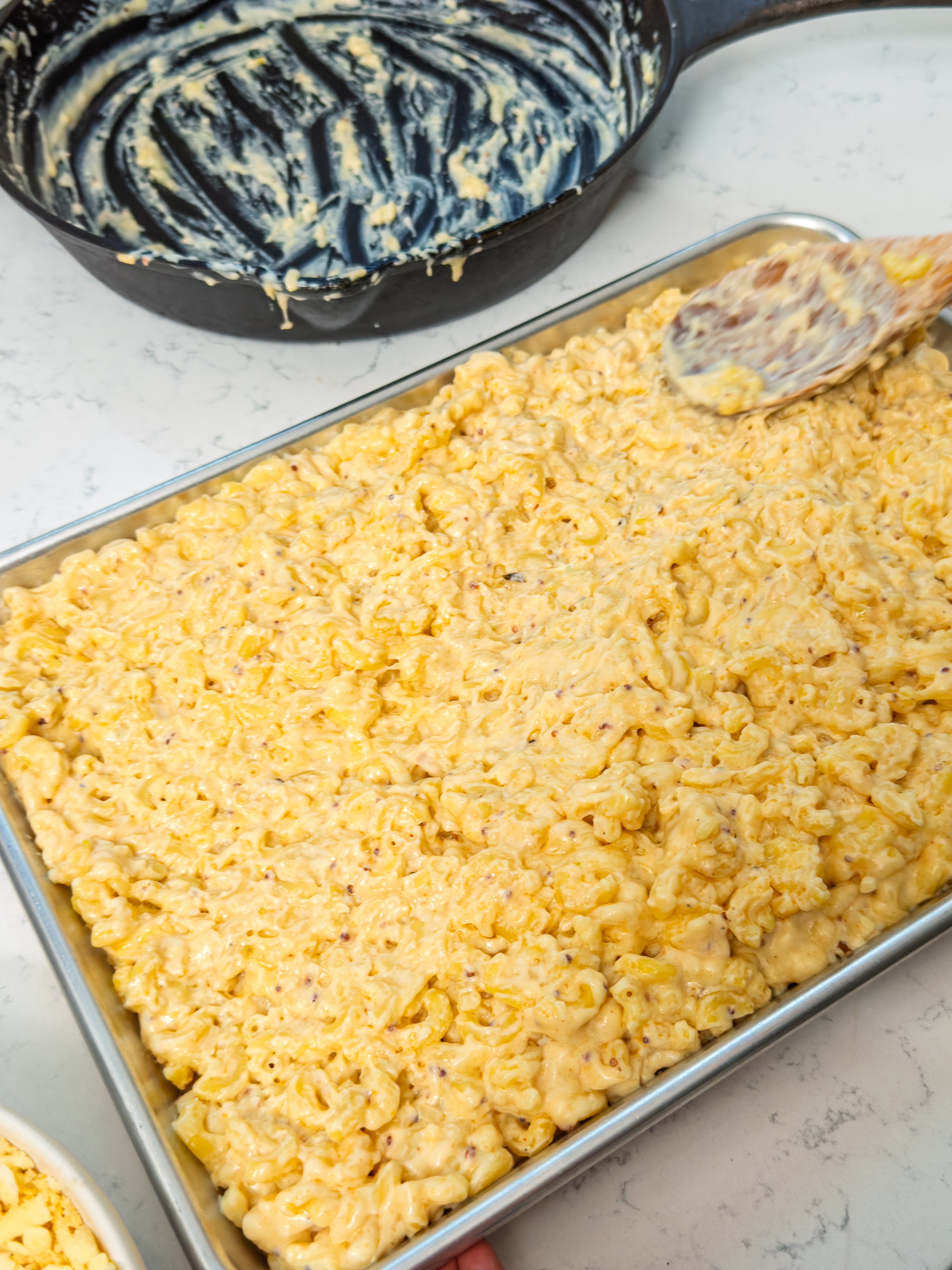 Baked macaroni and cheese in a rectangular pan, with a serving spoon and a pot with remnants of cheese sauce nearby