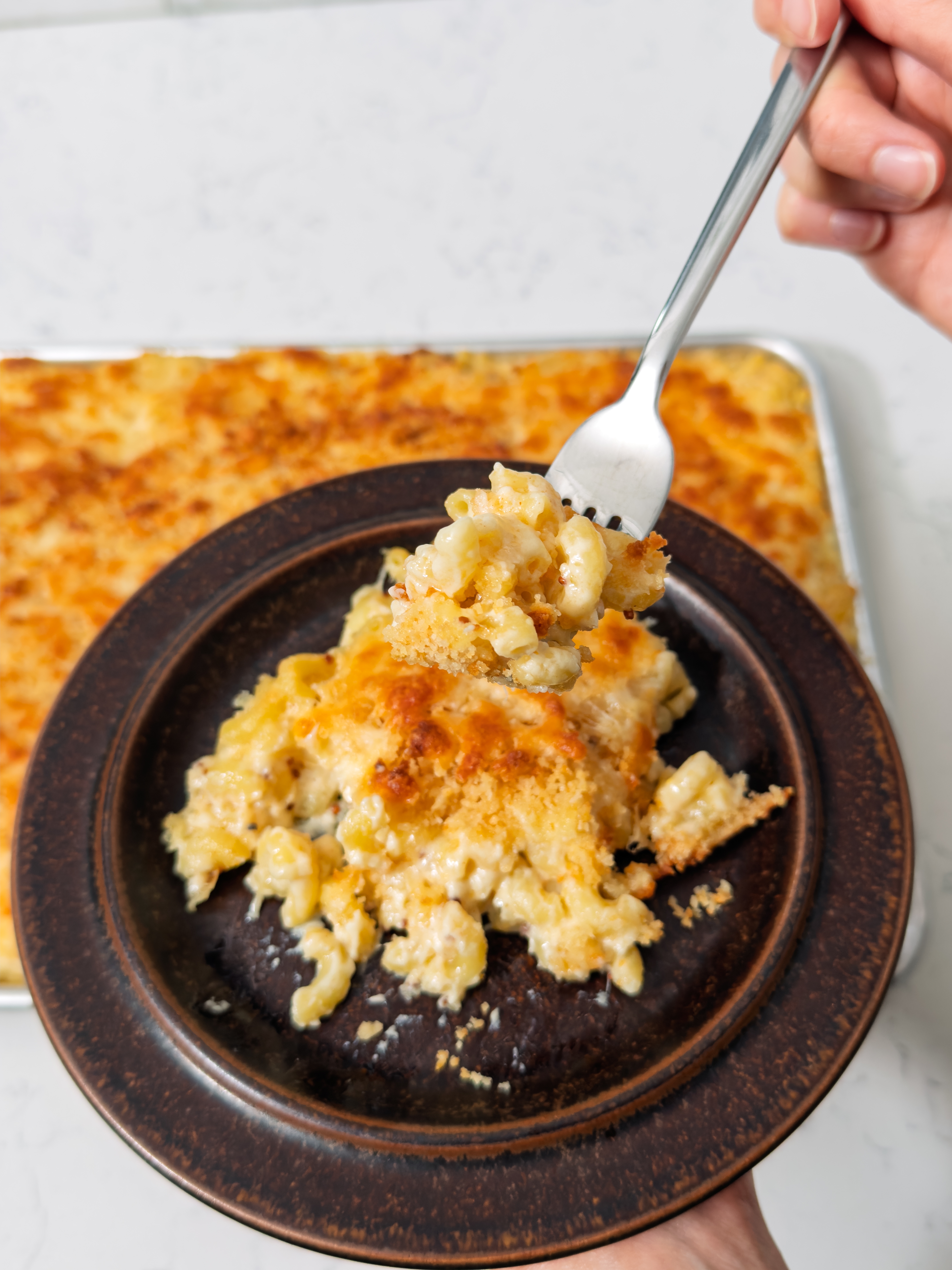 A fork lifts a creamy, baked macaroni and cheese serving from a dark dish; a large pan of mac and cheese is visible in the background