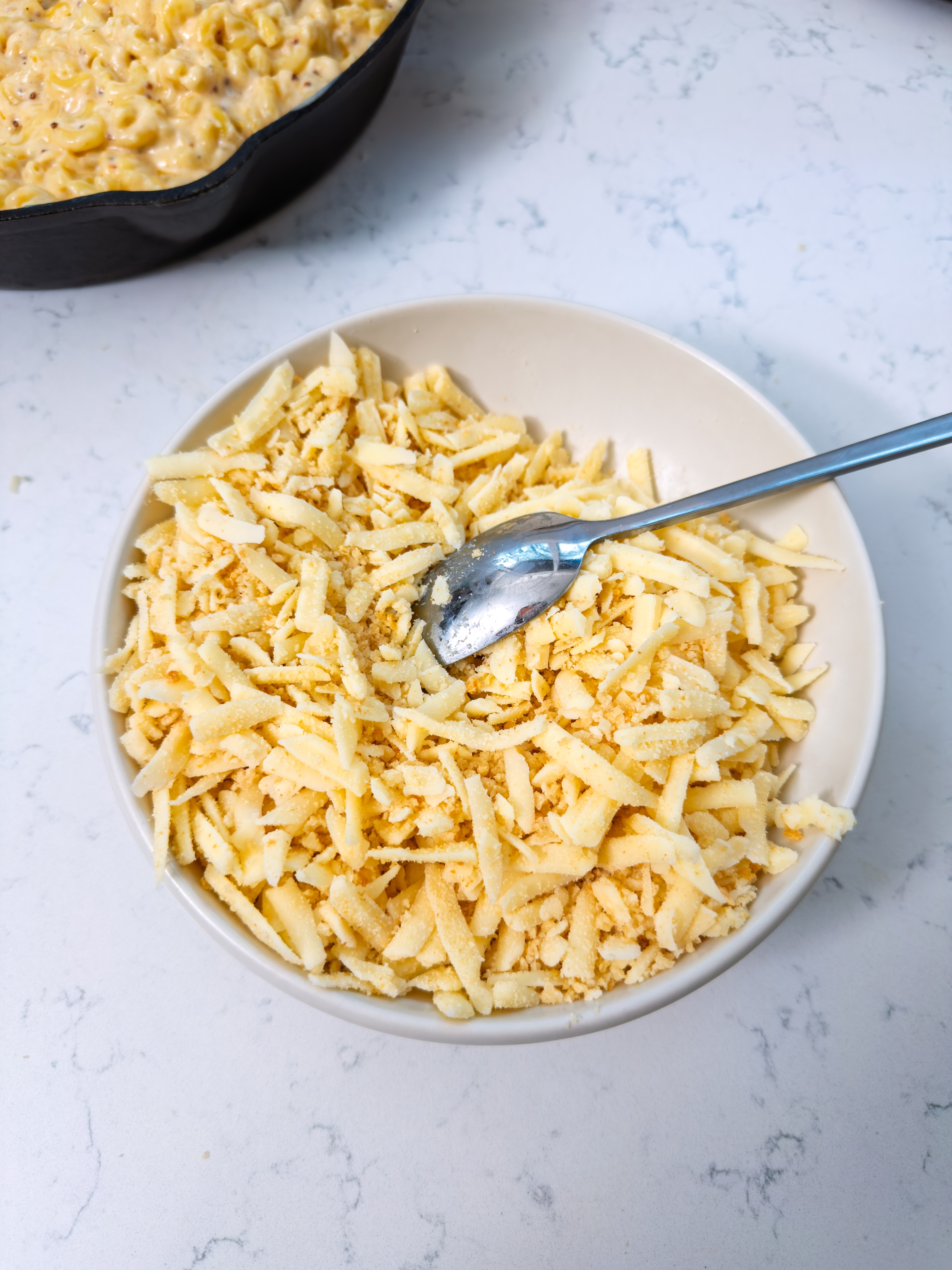 Bowl of grated cheese with a spoon on a white surface, with a skillet of mac and cheese in the background
