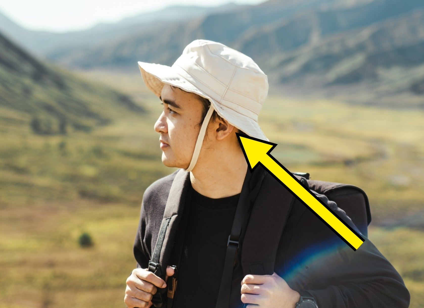 Person i en bredbremmet hatt og ryggsekk ser på et fjellandskap, kamera hengende fra nakken, foreslår eventyr eller fotturer