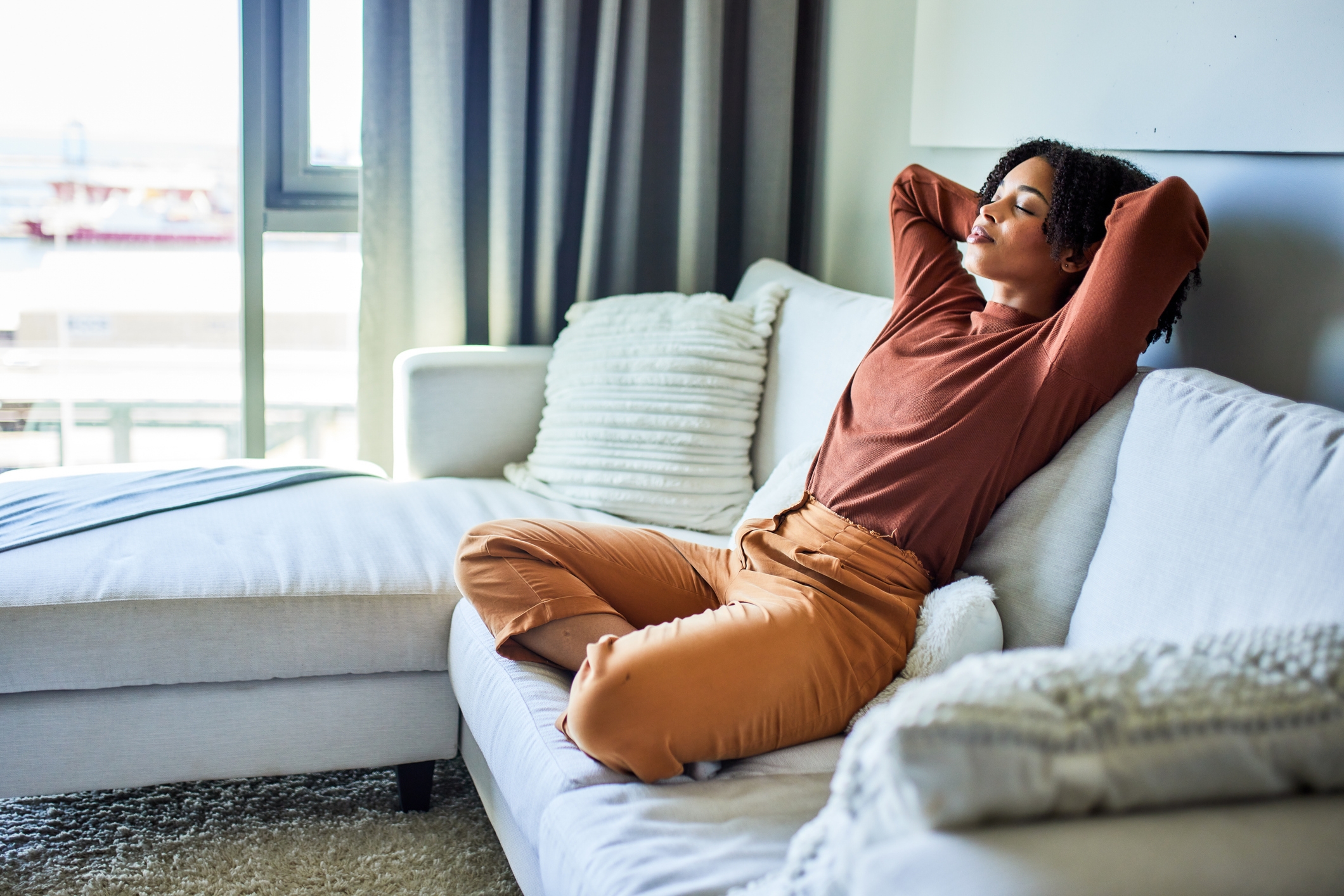 Person som slapper av på en sofa med lukkede øyne, armer bak hodet og bena i kors, virker fornøyd og veltilpas i en koselig stue