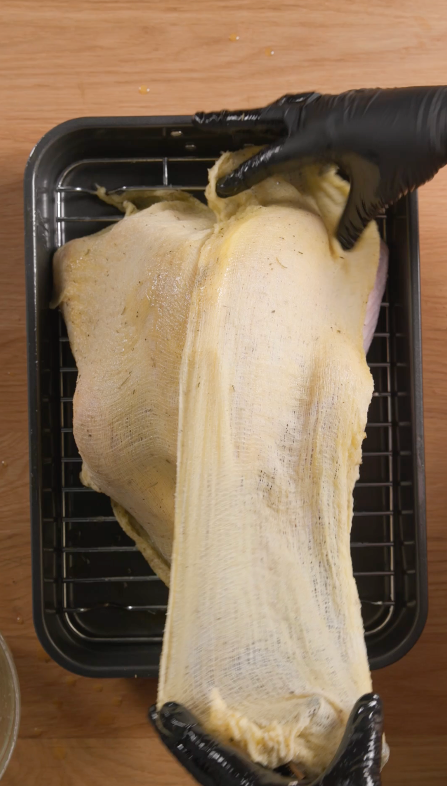 Person in black gloves wrapping seasoned raw meat in cheesecloth in a tray on a wooden surface