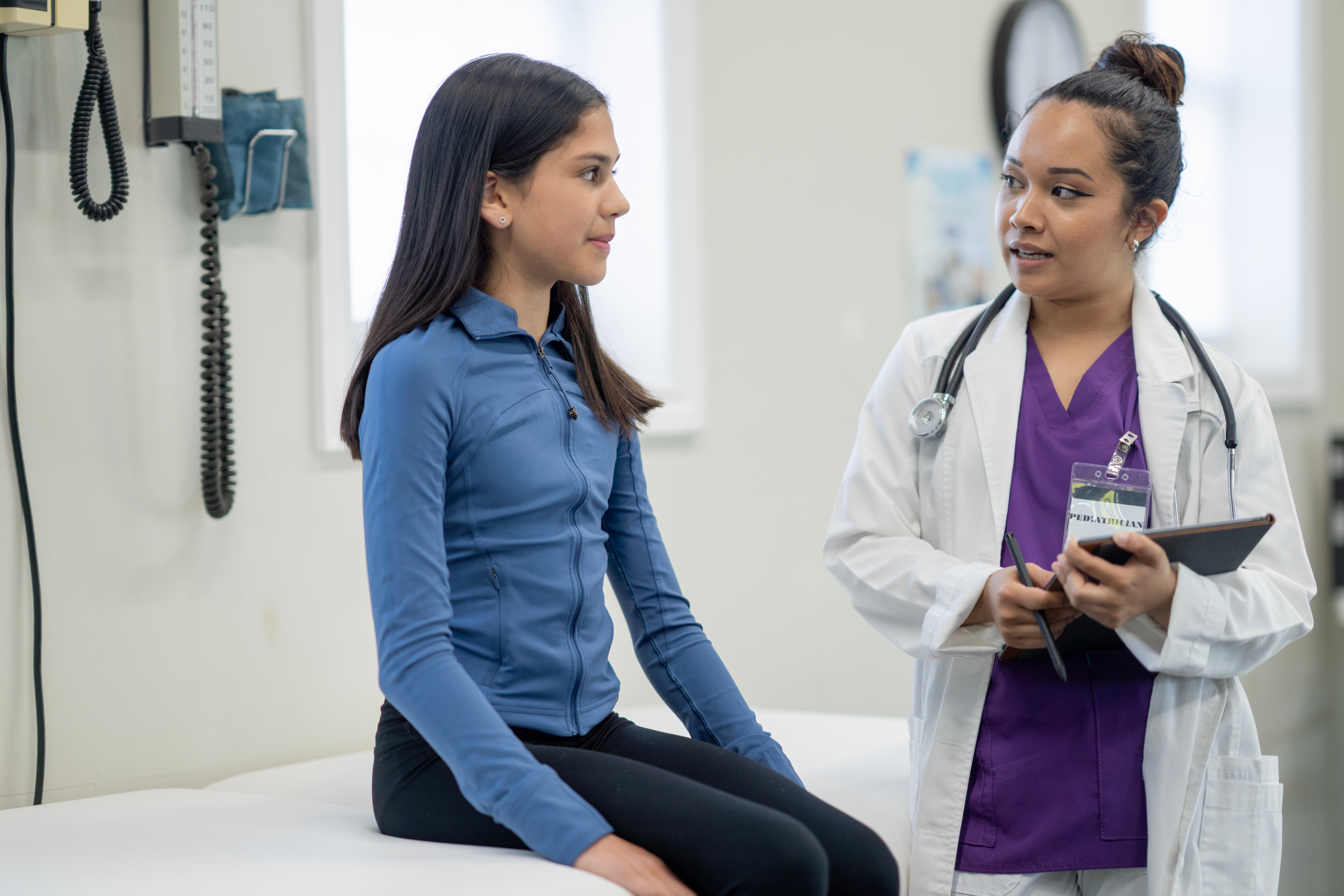 En ung kvinne sitter på et medisinsk undersøkelsesbord og snakker med en helsepersonell i lilla skrubber og hvit frakk og holder en nettbrett