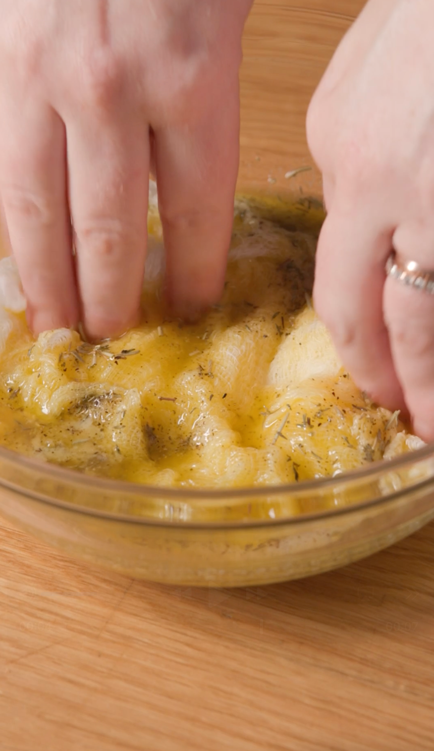 Hands marinate chicken in a glass bowl with oil and herbs