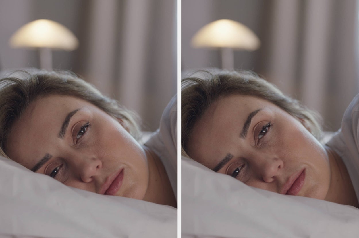 A person is lying in bed with an expression of tiredness or contemplation, resting their head on a pillow, with a lamp softly lit in the background