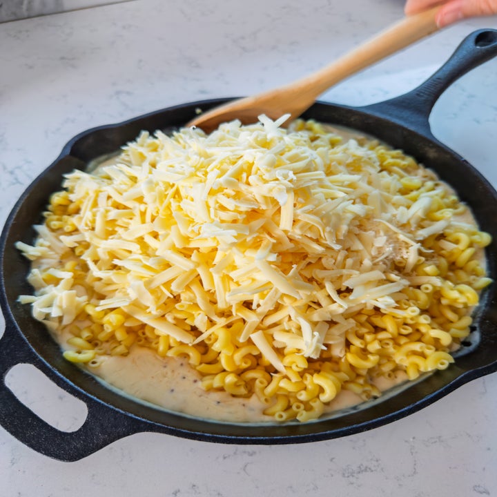 Pasta in a skillet mixed with creamy sauce and topped with a large heap of shredded cheese, being stirred with a wooden spoon