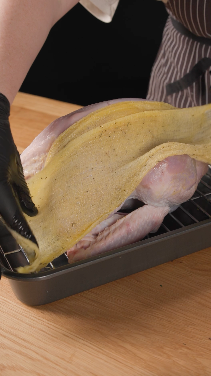 A person places a large sheet of seasoned dough over a raw turkey on a roasting rack, preparing it for cooking