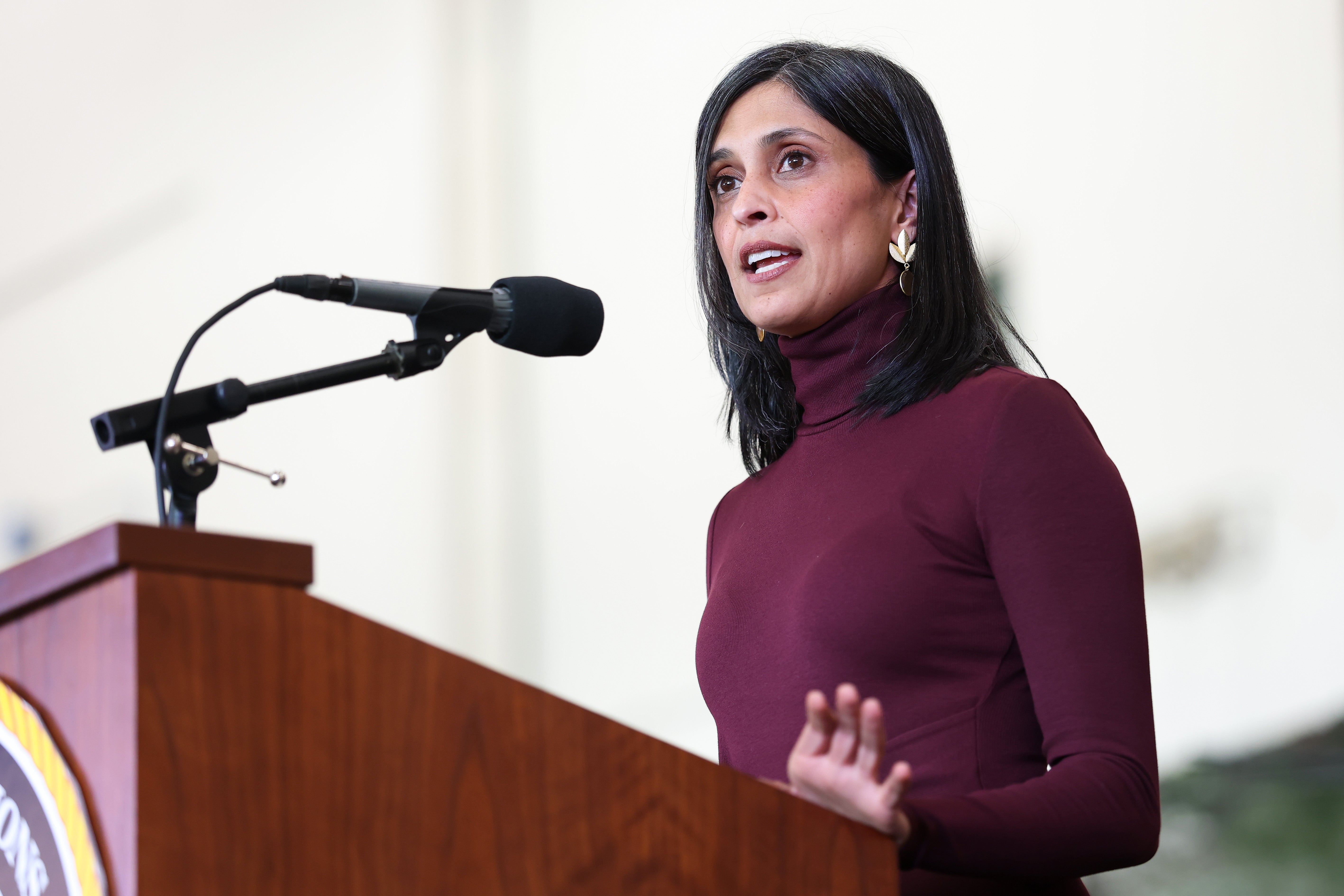 Person speaking at a podium with a microphone, wearing a fitted, high-neck top and statement earrings, gesturing with one hand