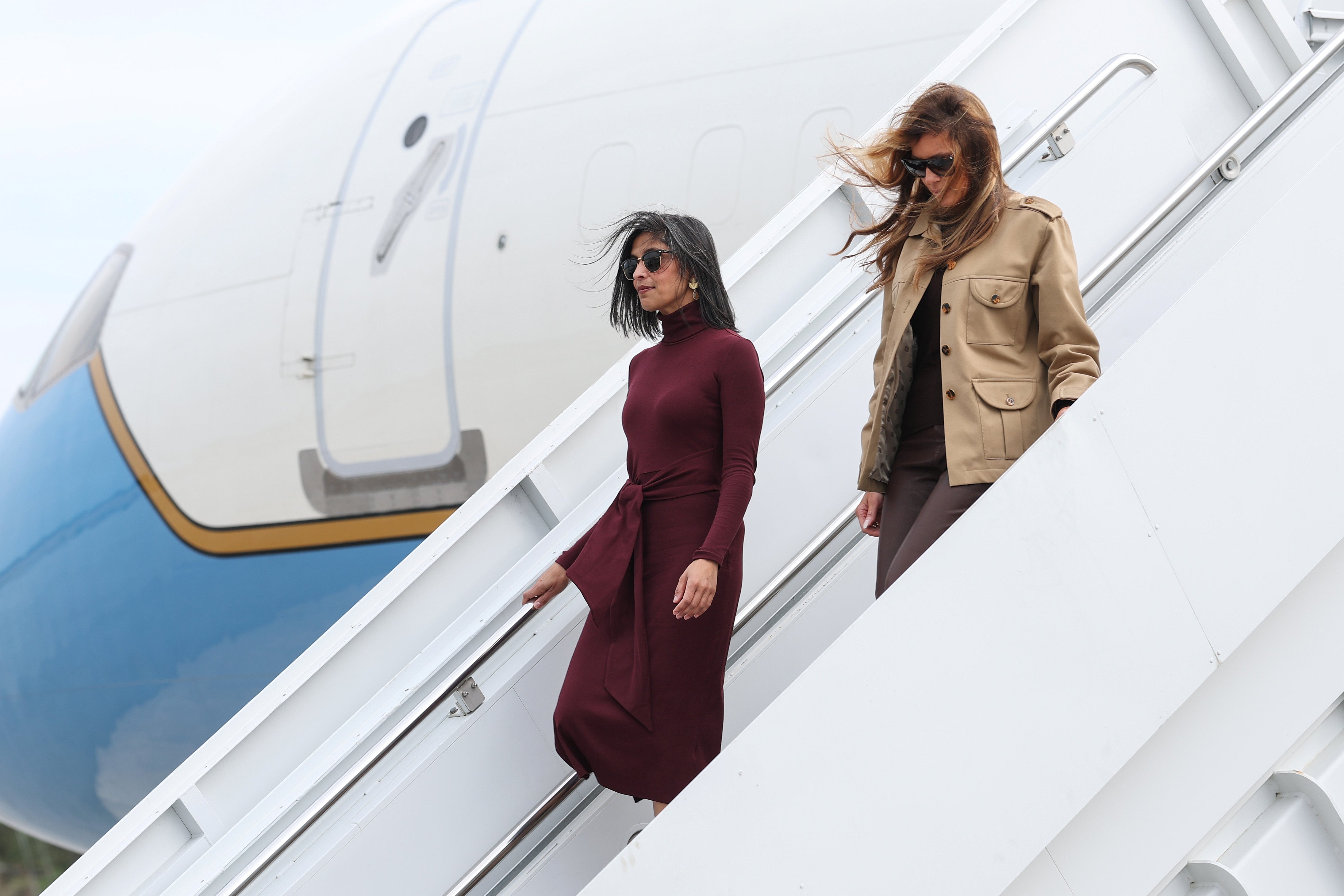 Two women descend airplane stairs, both wearing sunglasses. One wears a long-sleeved dress, the other a jacket and pants
