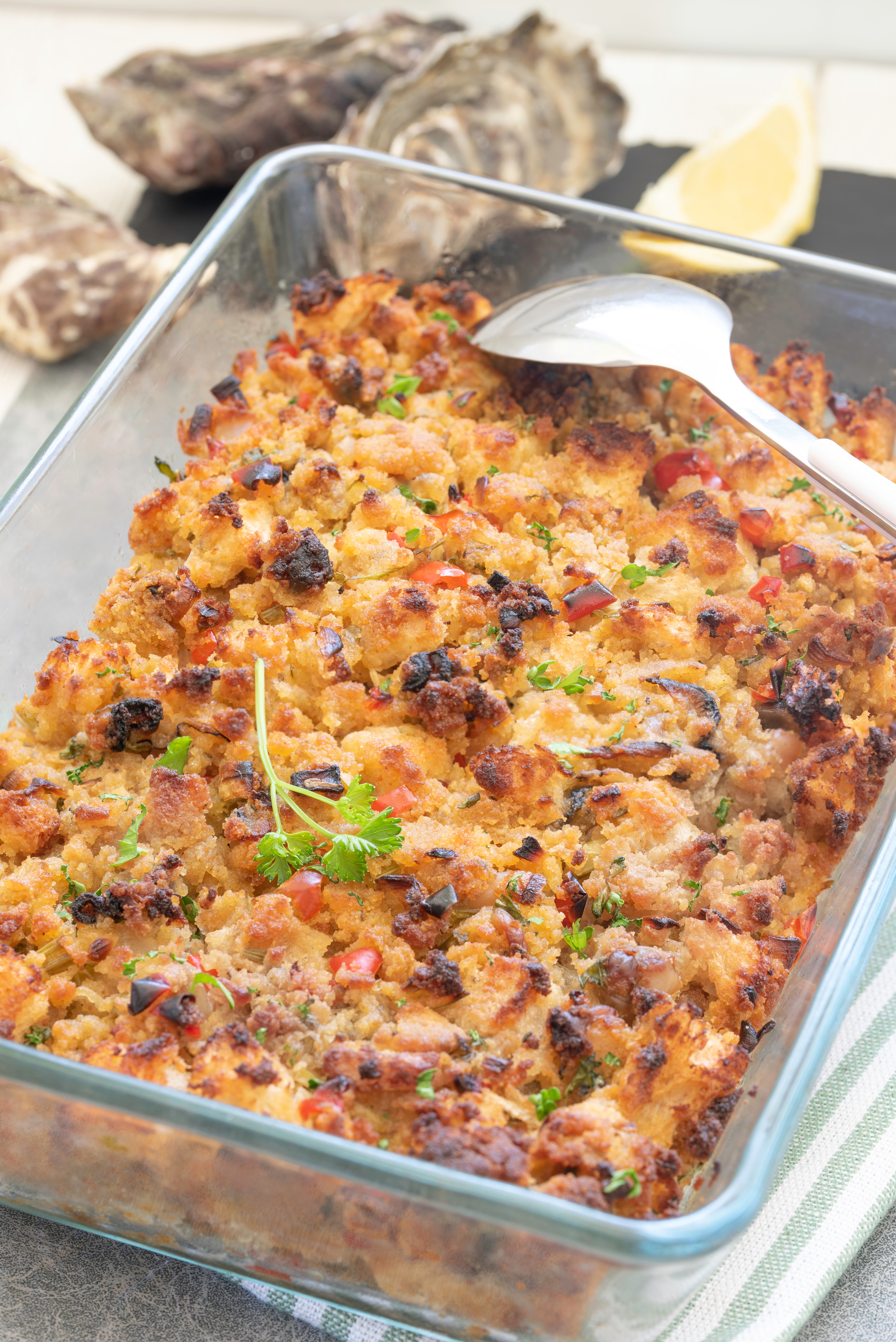 Baked seafood stuffing in a glass dish, topped with herbs, surrounded by lemon wedges and oysters