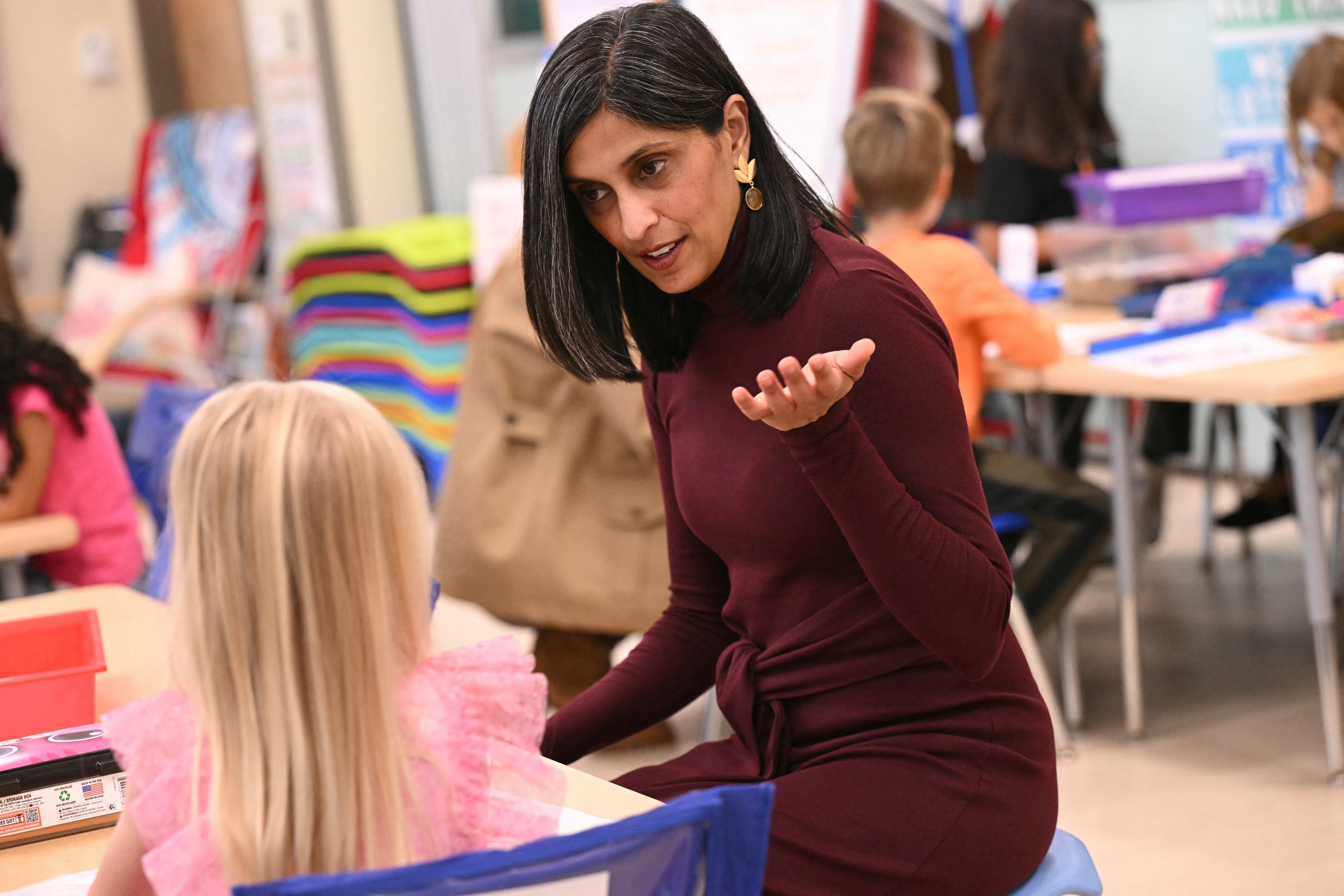 A woman in a dress engages with a child in a classroom setting, both seated at tables with other children in the background
