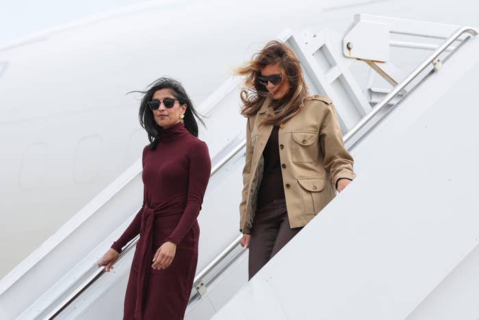 Two women walking down an airplane staircase; one in a maroon dress, the other in a tan jacket and pants, both wearing sunglasses