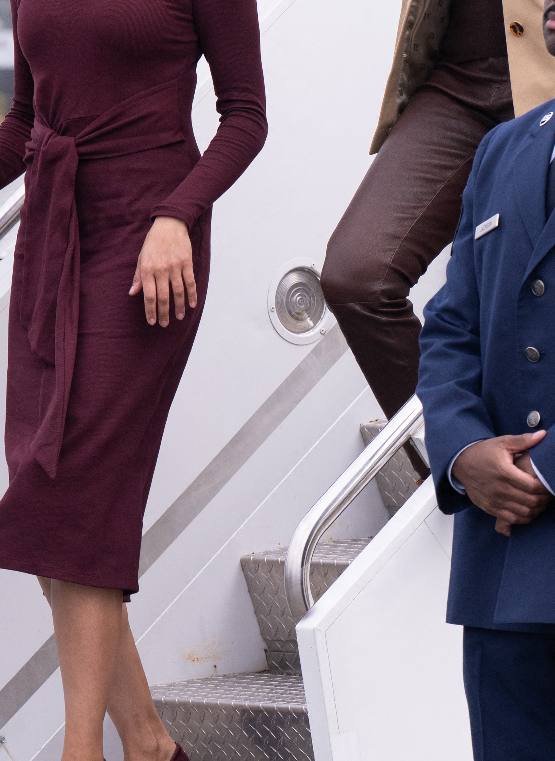 Two people in stylish attire descending airplane steps, surrounded by uniformed personnel on an airport tarmac