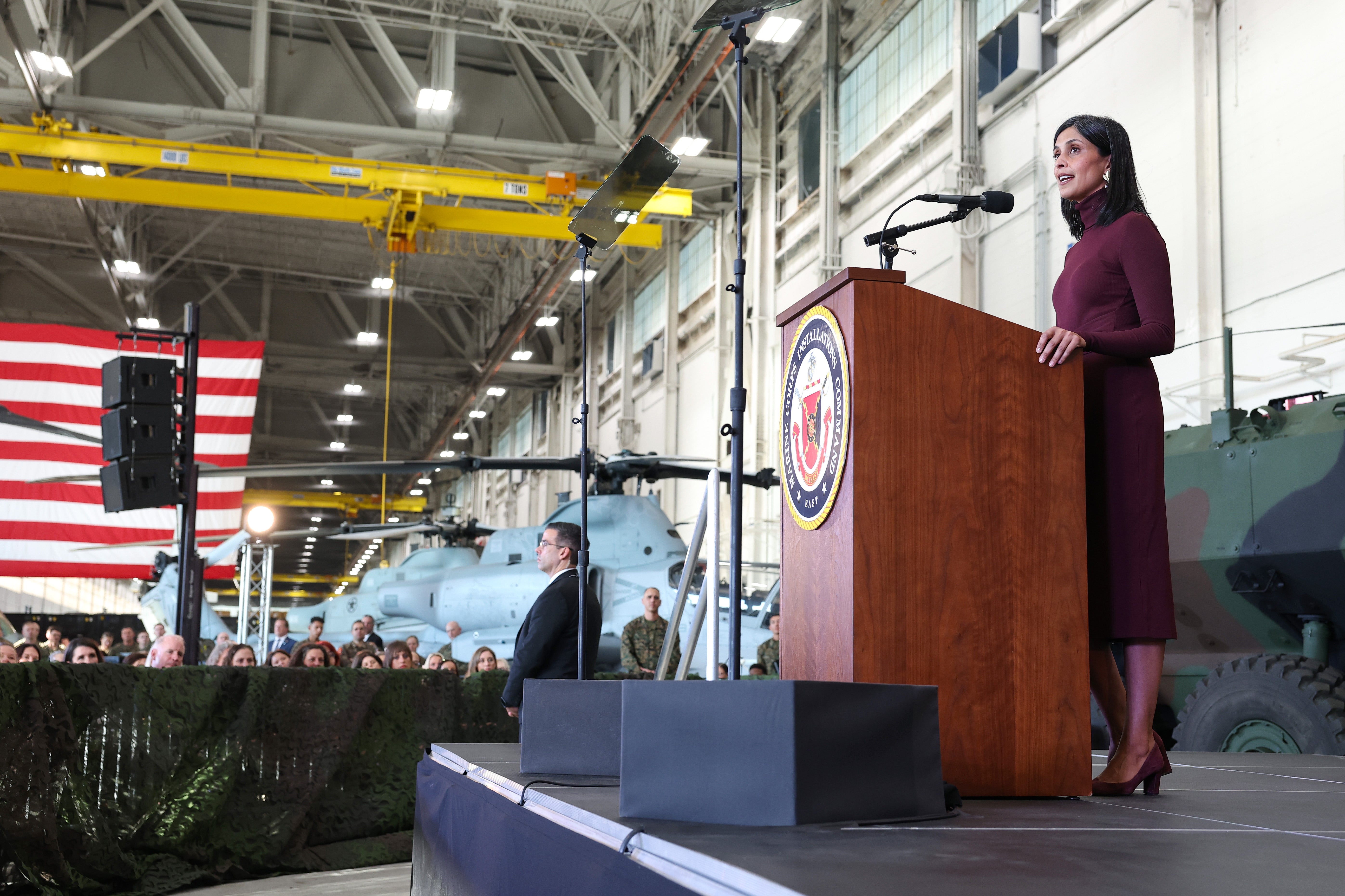 A woman speaks at a podium inside a hangar with military aircraft in the background. An audience and an American flag are visible