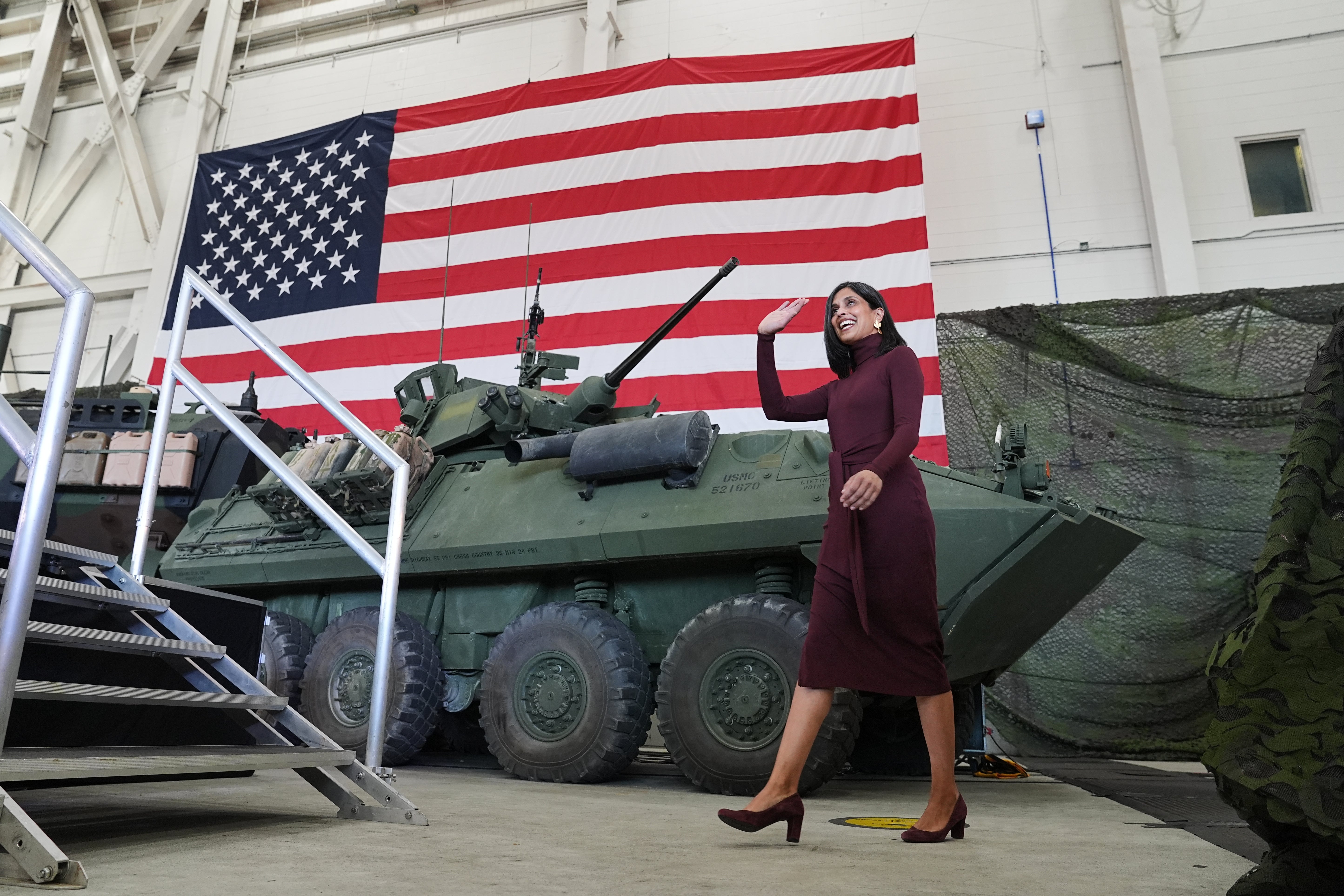 A woman in a long dress and heels waves while walking past a military vehicle inside a hangar with a large U.S. flag in the background