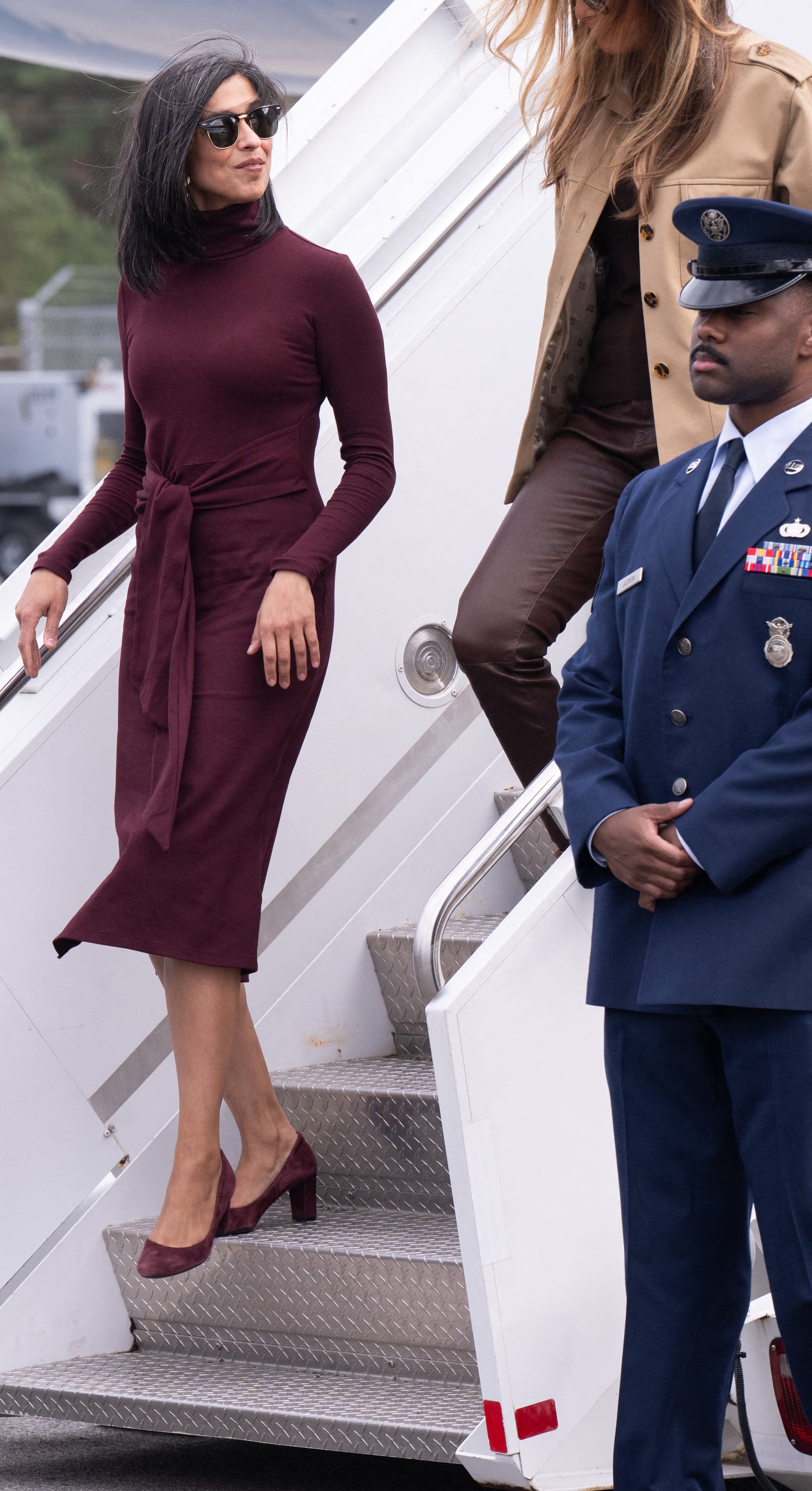 Two women in stylish outfits descend airplane steps, surrounded by three uniformed guards on a tarmac