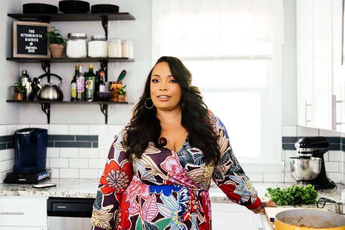 A person stands in a modern kitchen wearing a floral dress, with shelves of spices and utensils behind them, seemingly engaged in cooking or food preparation