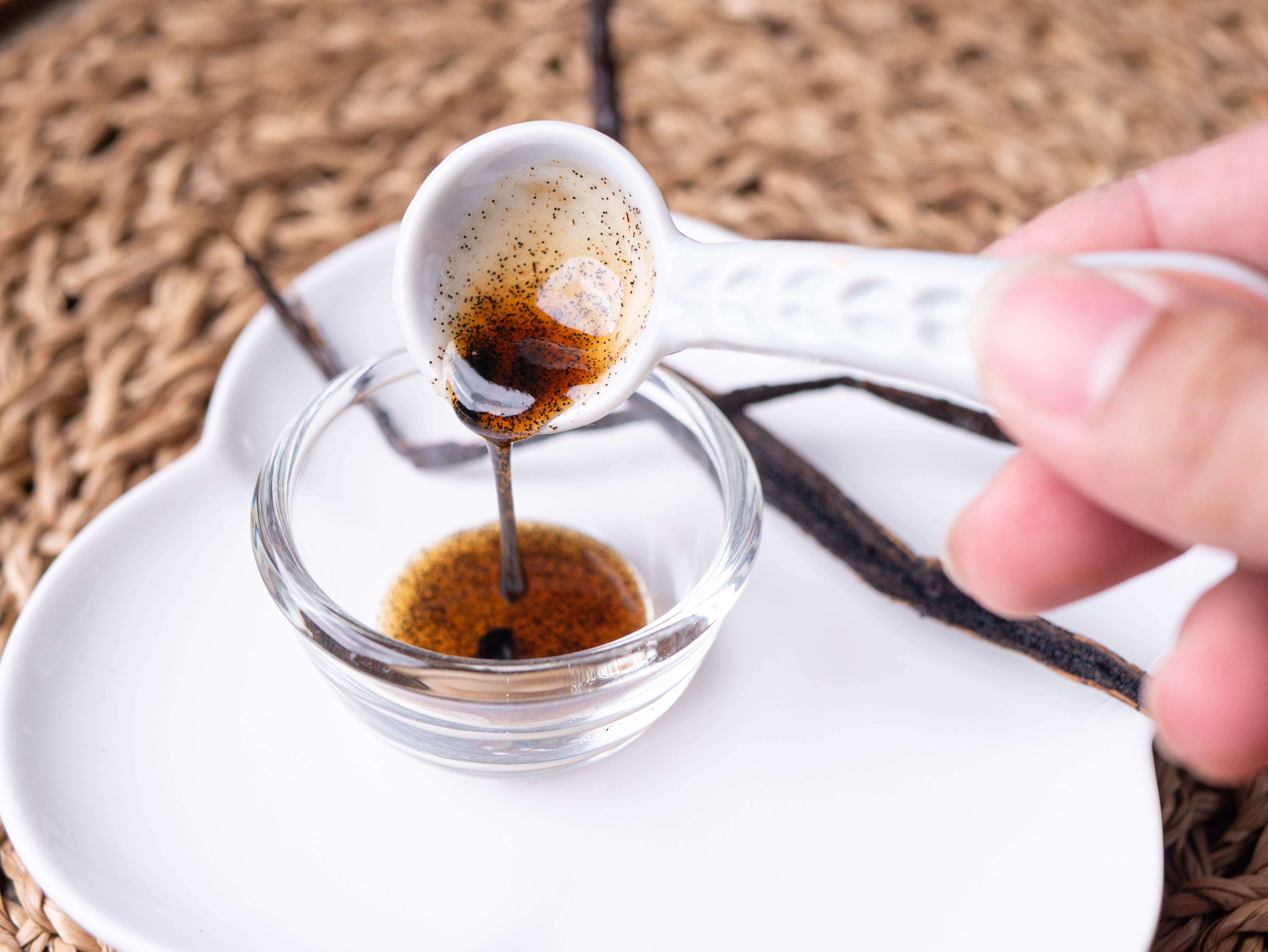 Vanilla extract being poured from a spoon into a small glass bowl, with vanilla pods in the background