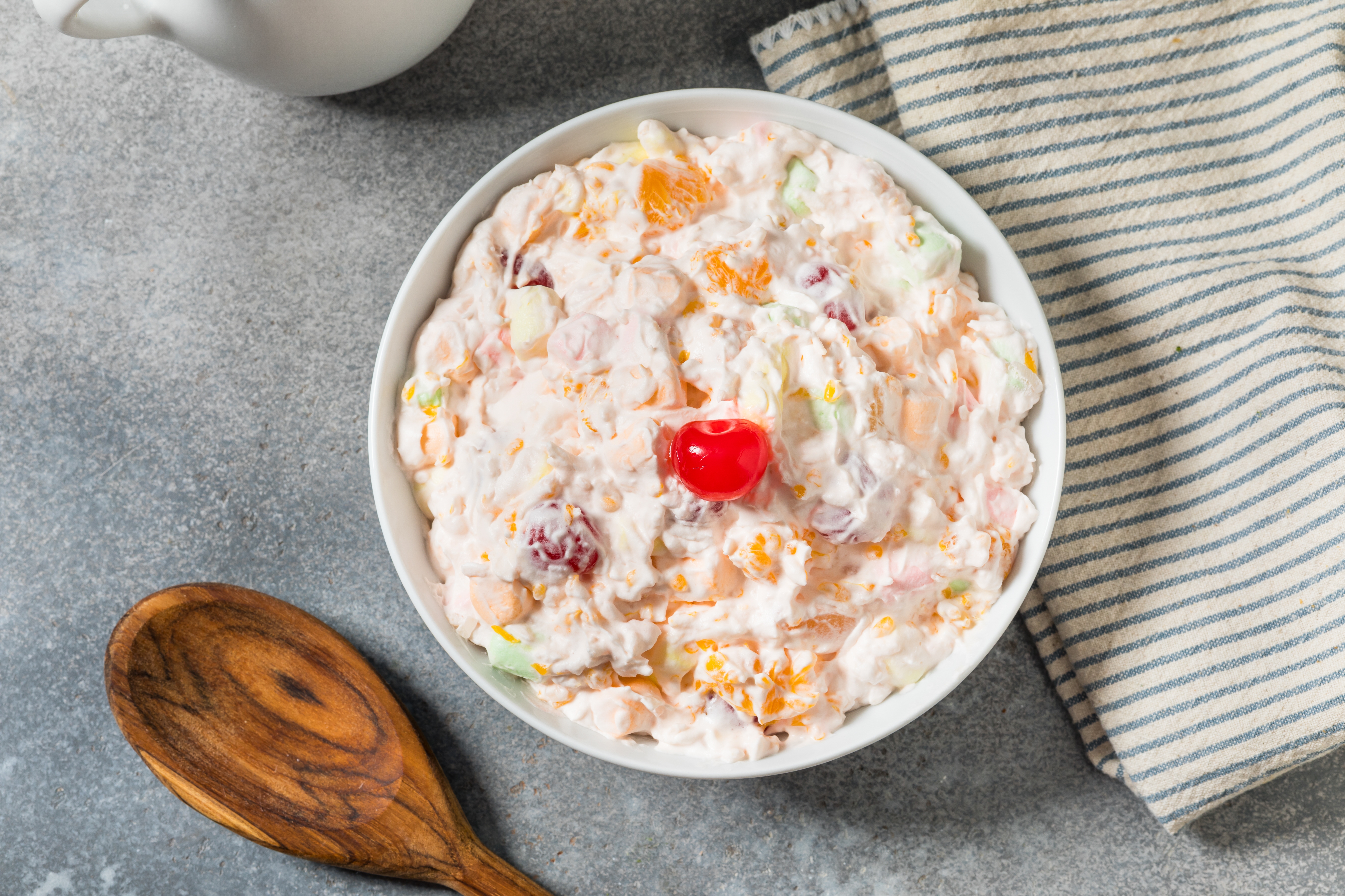 A bowl of fruit salad with whipped cream, topped with a cherry, on a table beside a wooden spoon and striped cloth