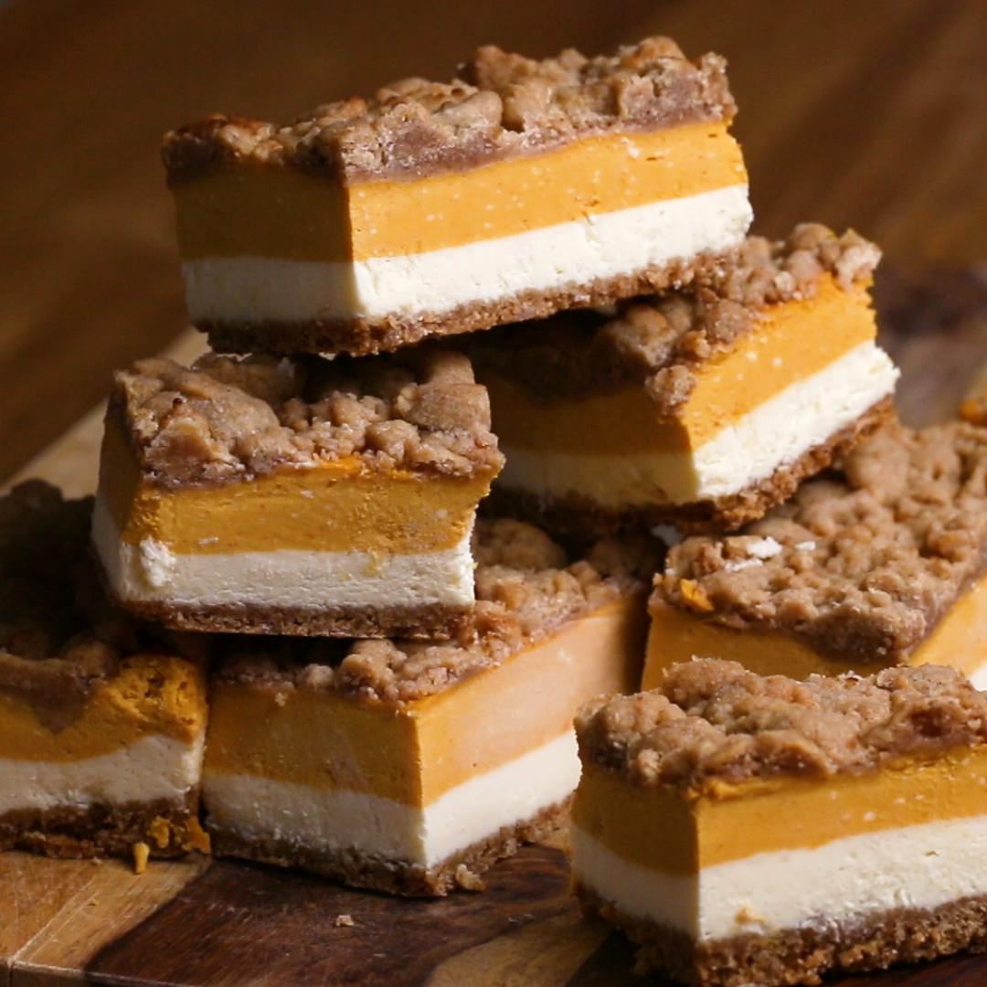 Layered dessert bars with creamy pumpkin, cheesecake, and crumb topping, stacked on a wooden board