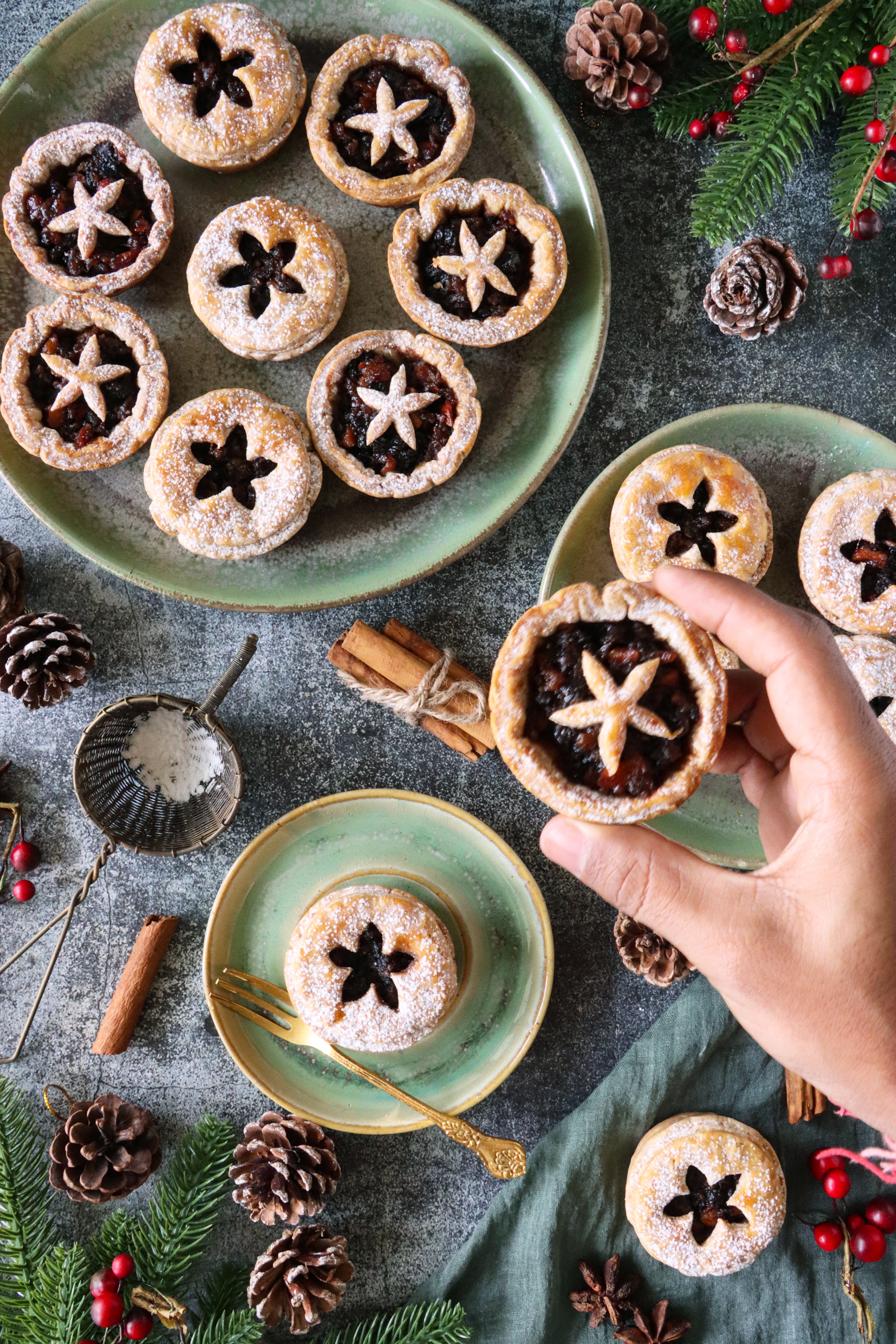 A hand holds a mince pie with a star-shaped crust on a table set with more mince pies, pine cones, and festive greenery