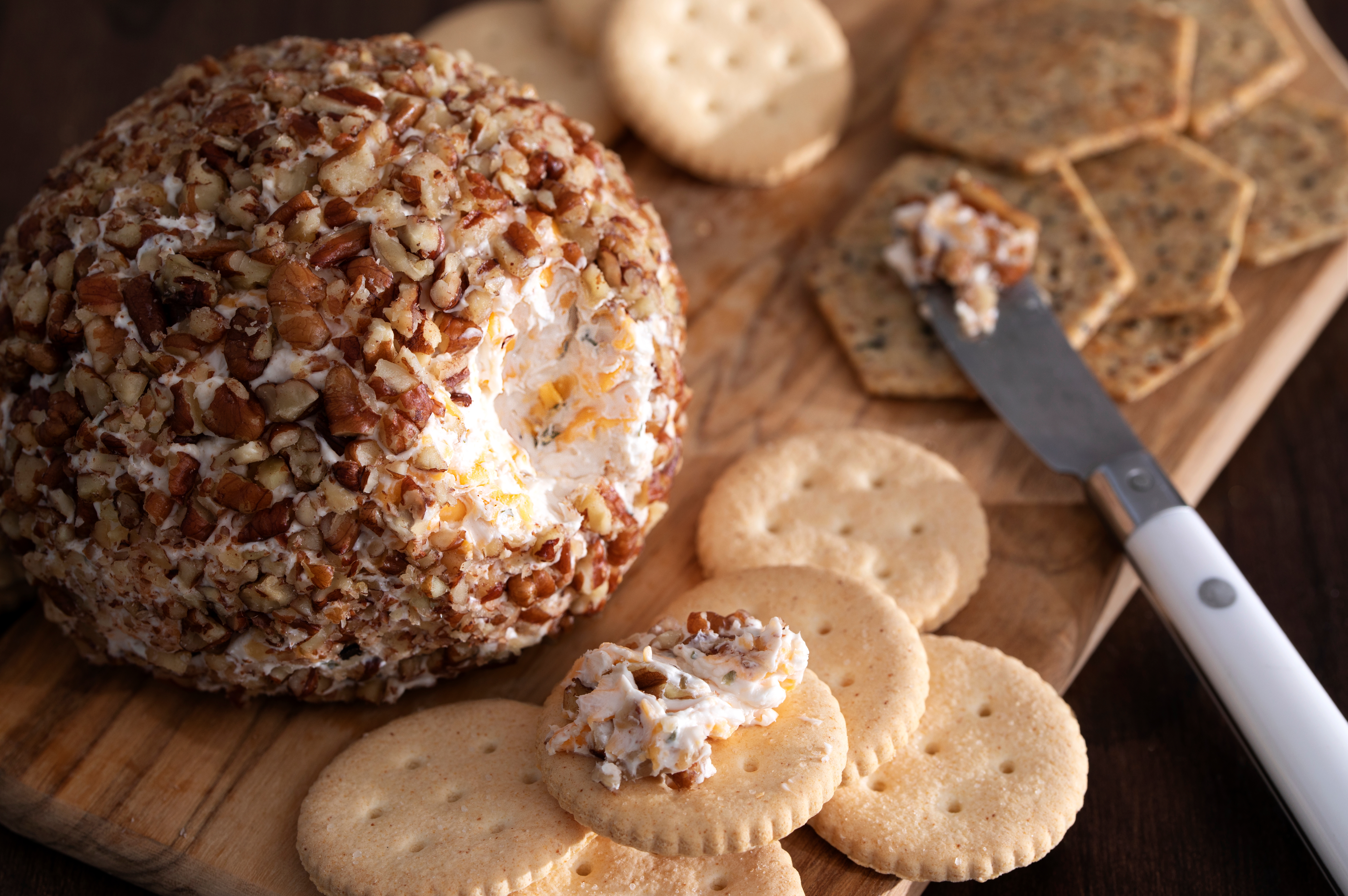 Cheese ball coated in chopped nuts on a wooden board, surrounded by assorted crackers, with a knife spreading cheese