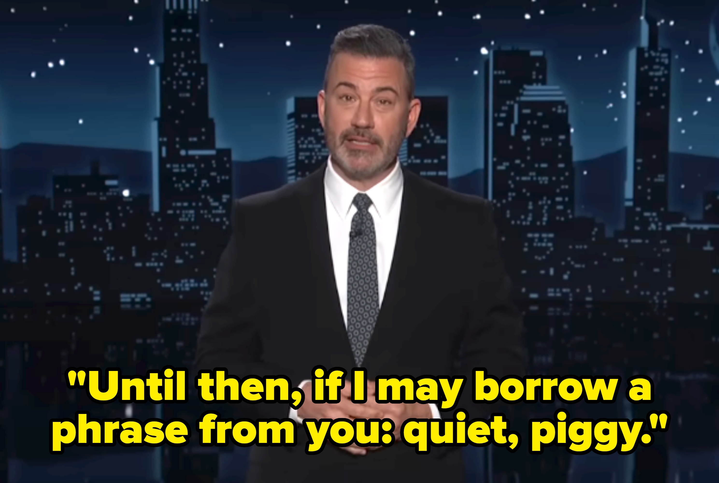 A person in a suit on a night-time talk show set with a cityscape backdrop