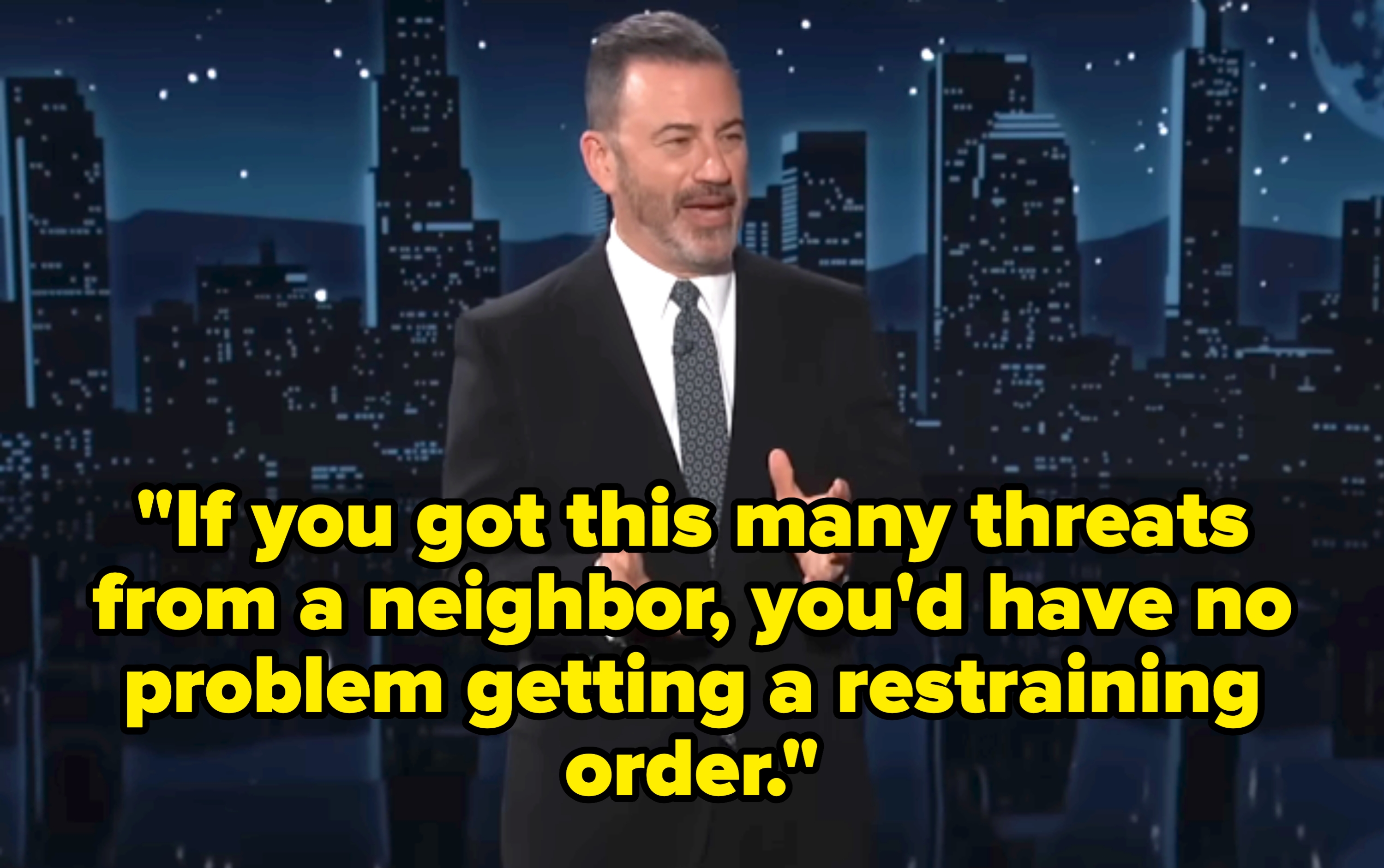 A man in a suit and tie stands against a city skyline backdrop, speaking animatedly