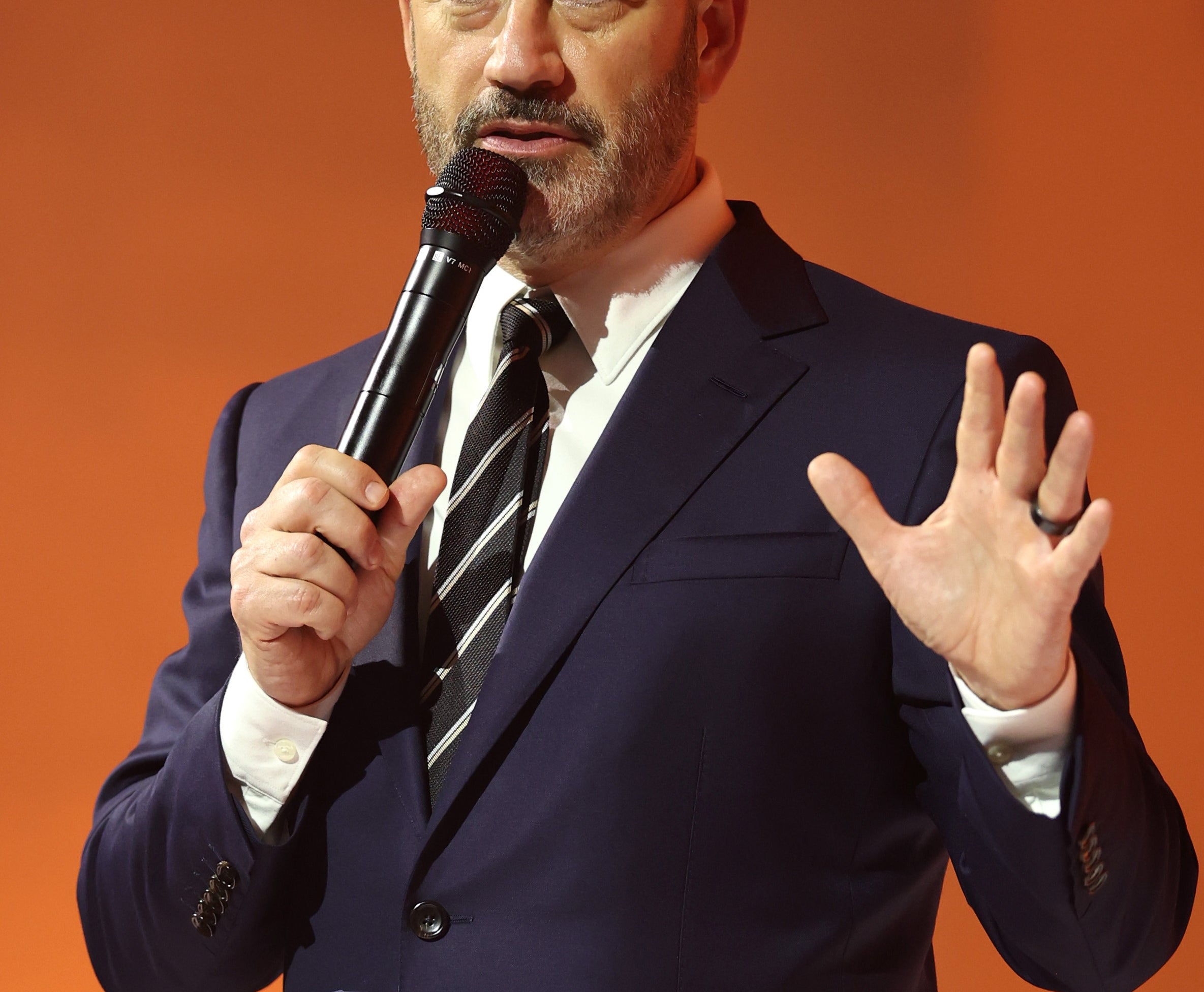 Person in a formal suit holds a microphone, speaking onstage