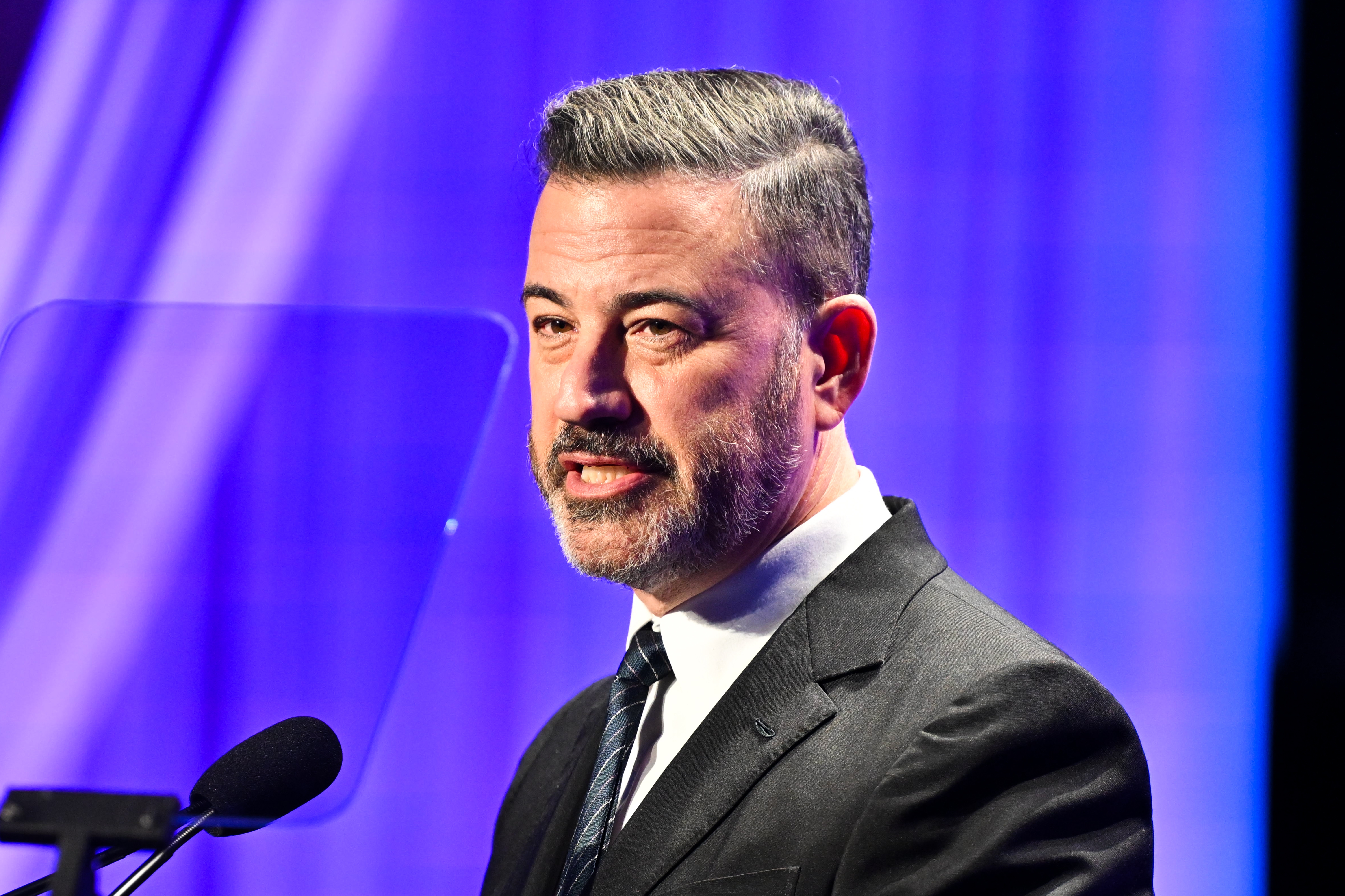 Man in a suit speaking at a podium during an event, with a blue-lit background