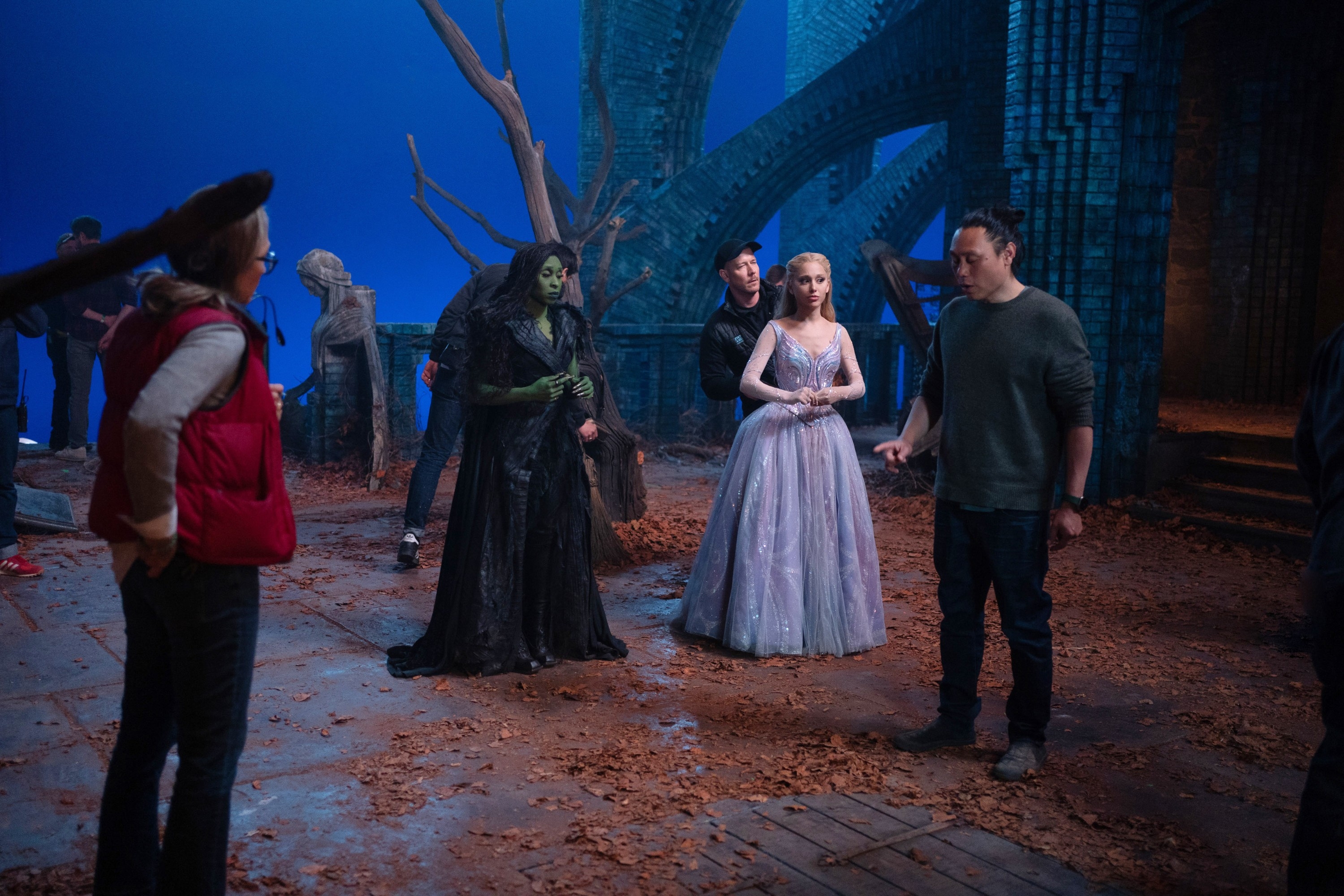 People on a film set with fantastical costumes. One wears a long black outfit and green makeup, another a sparkly gown. Crew members are seen around