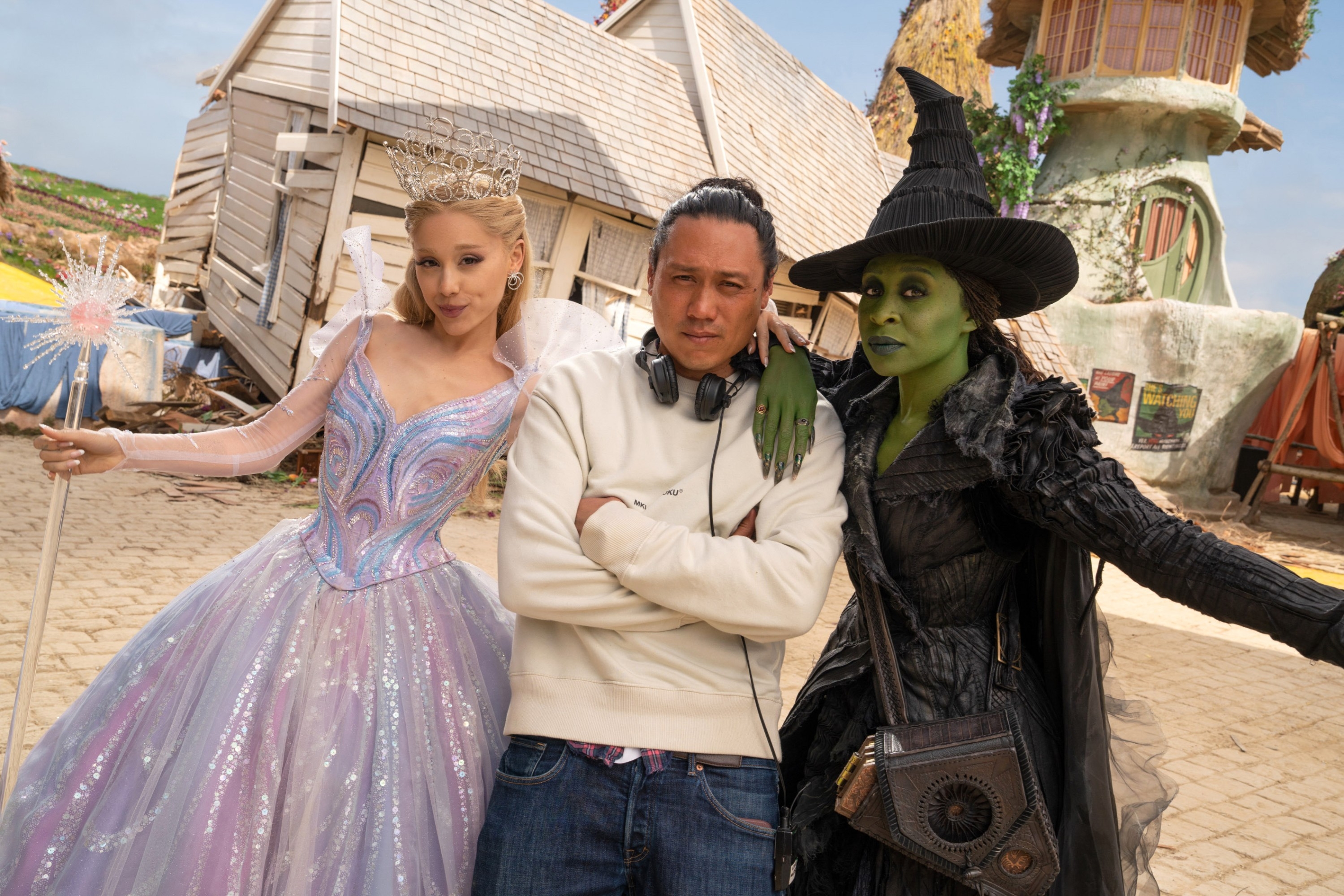 Ariana Grande and Cynthia Erivo in elaborate fairy tale costumes pose with Jon M. Chu on a whimsical set