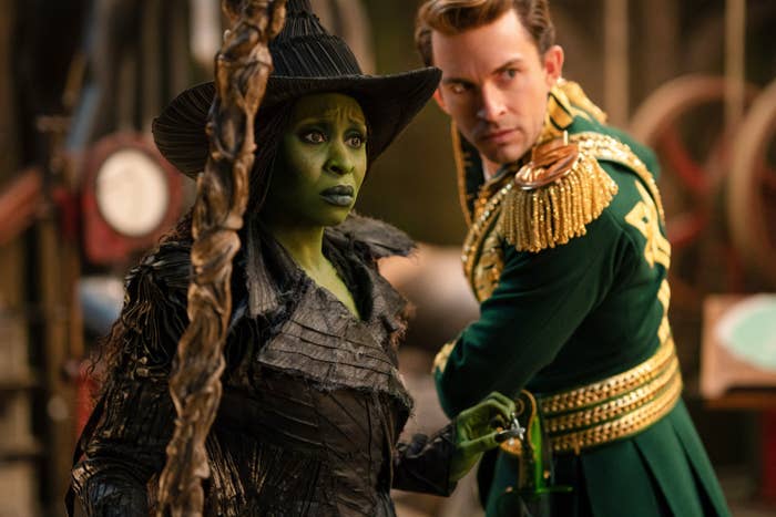 Cynthia Erivo and Jonathan Bailey in theatrical costumes. One is dressed as a witch with a textured hat, while the other wears a military-style outfit with gold accents