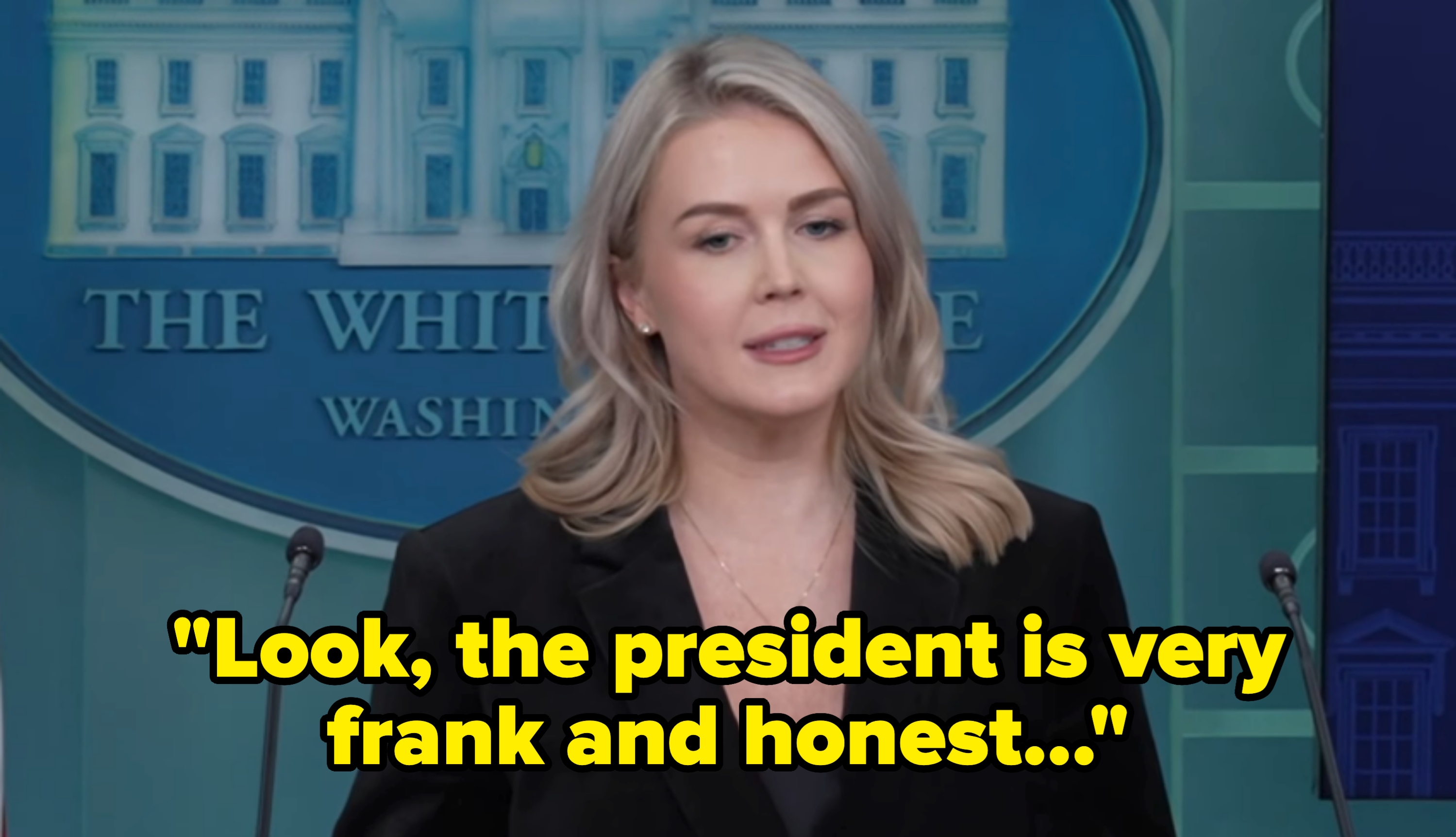A person stands at a podium in a press room, wearing a black blazer with a necklace, in front of a White House sign