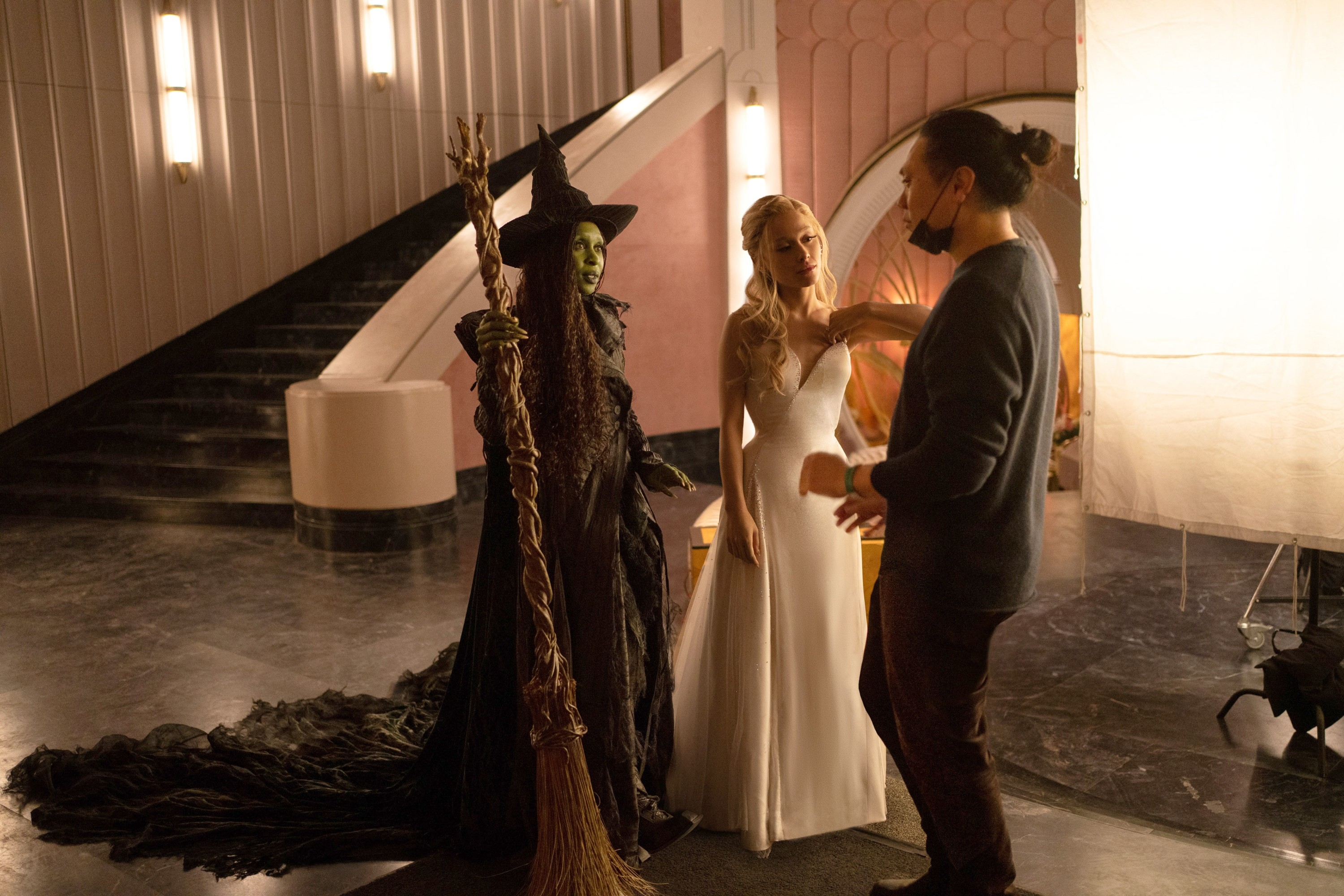 Cynthia Erivo and Ariana Grande in costumes, one as a witch with a hat and one in a white gown, are on set with Jon M. Chu for a possible film or theater production