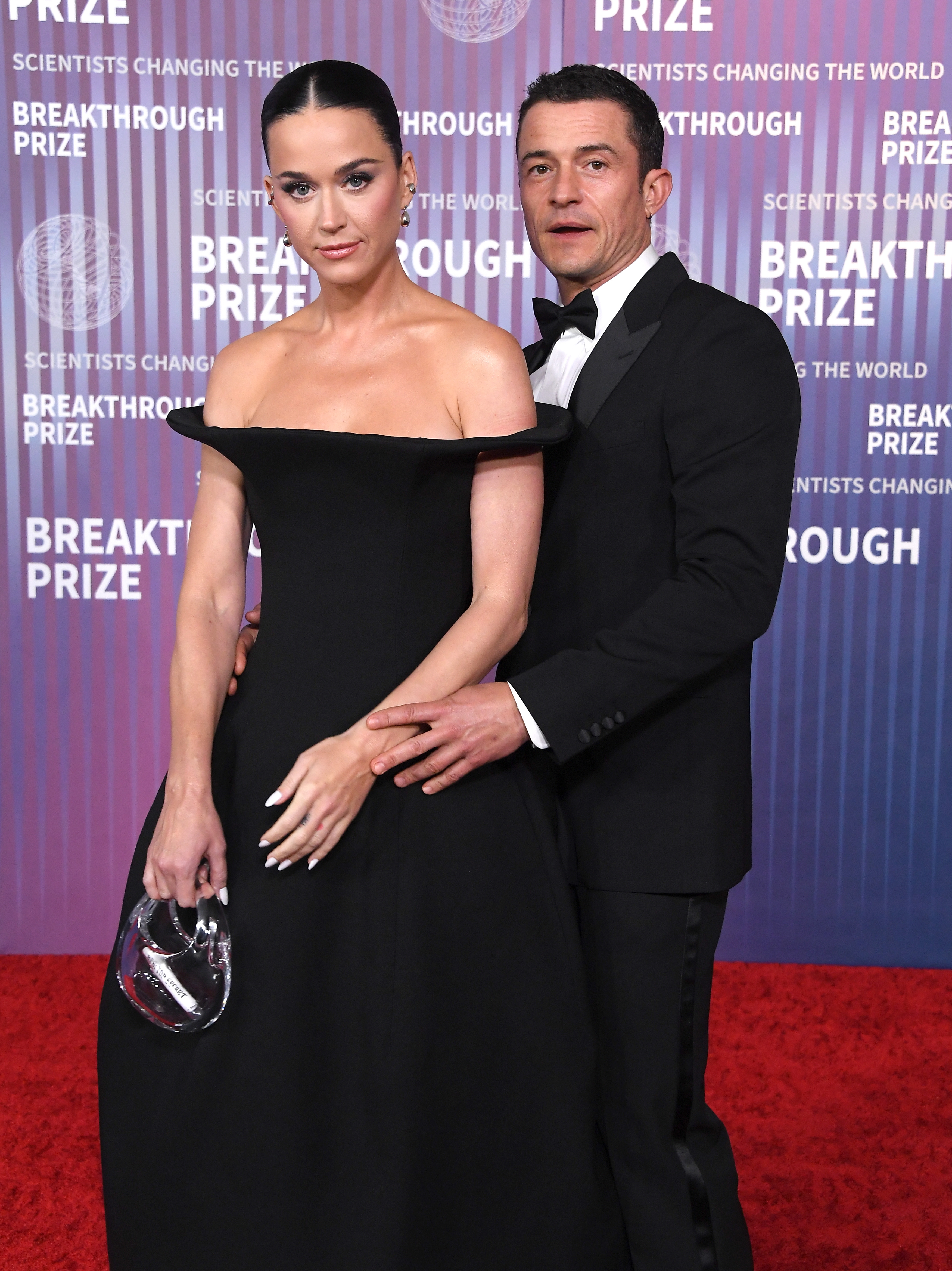Katy Perry and Orlando Bloom poses on the red carpet at the Breakthrough Prize event; the woman wears an elegant off-shoulder gown, and the man wears a classic tuxedo