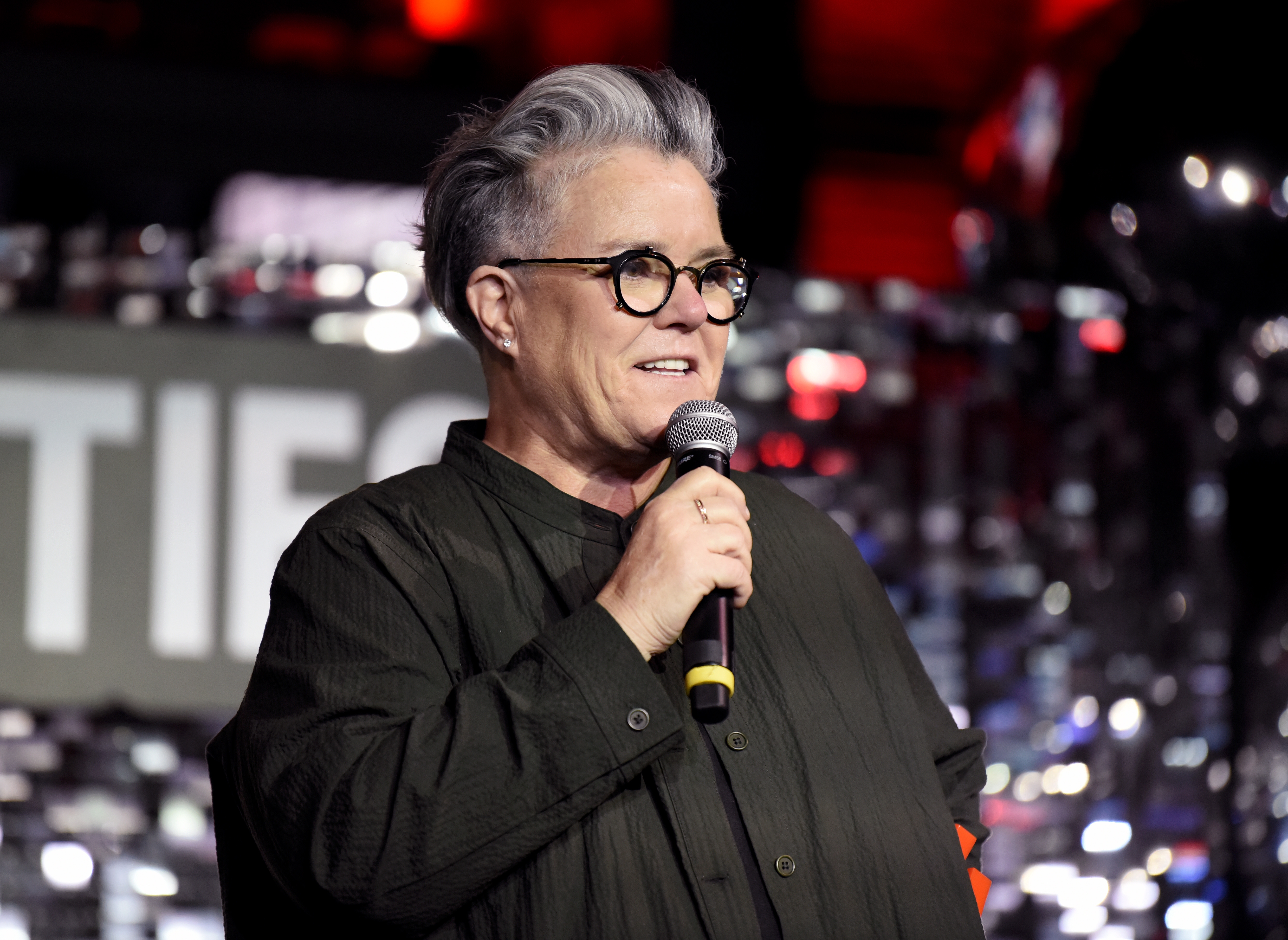 Rosie O'Donnell with short hair and glasses on stage, holding a microphone, wearing a loose, shirt, with a blurry background of event signage
