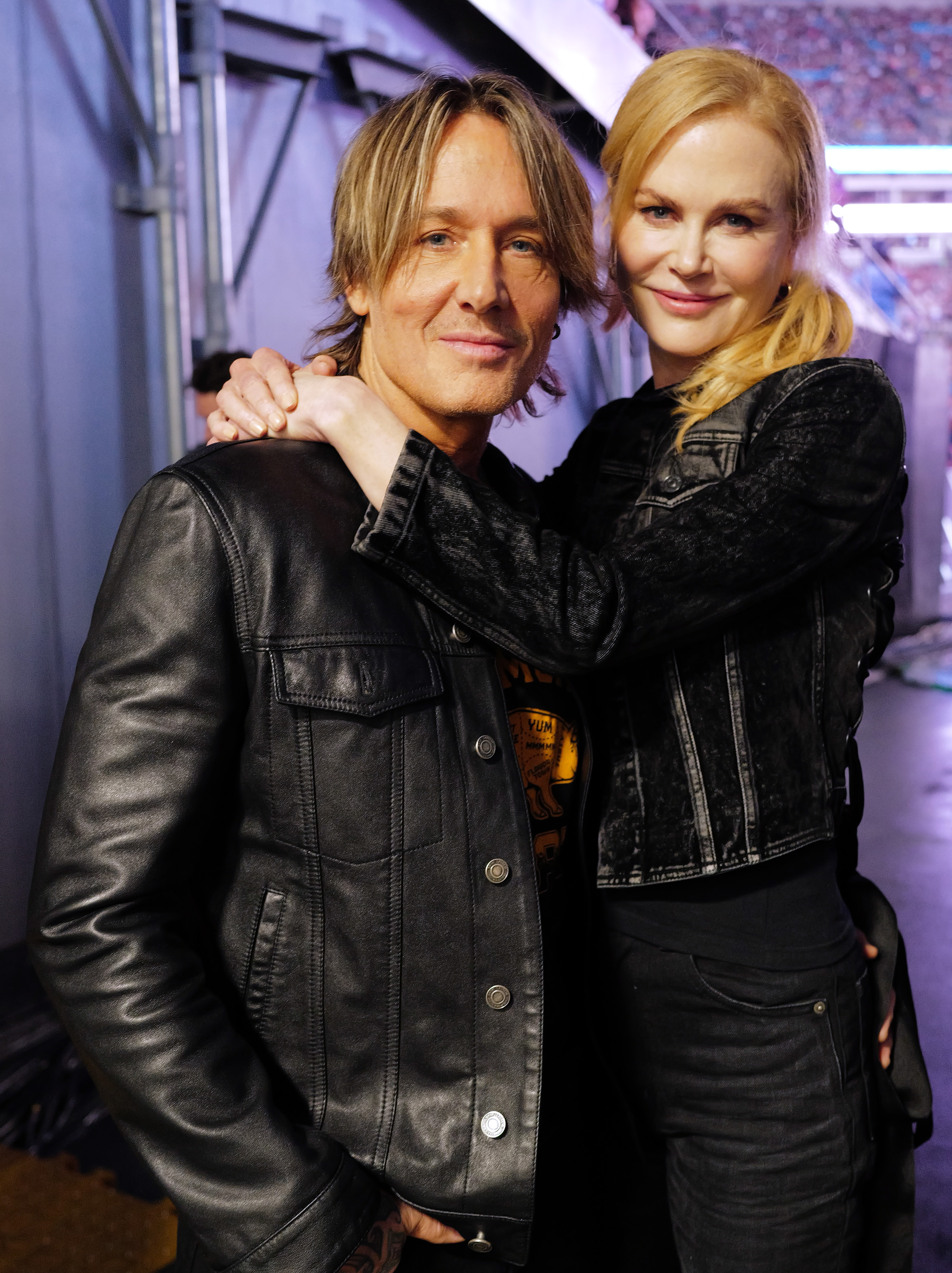 Two people in black leather jackets share a warm, friendly pose. One has shoulder-length hair, and the other's hair is styled in a ponytail