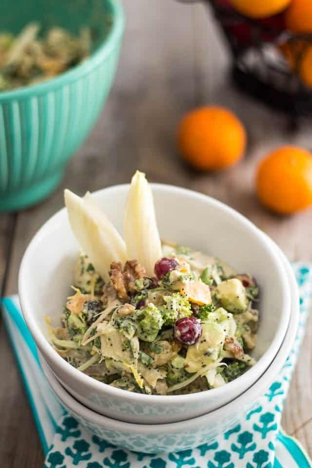 A salad in a white bowl with broccoli, walnuts, creamy dressing, and decorative endive leaves. Oranges and a green bowl are in the background