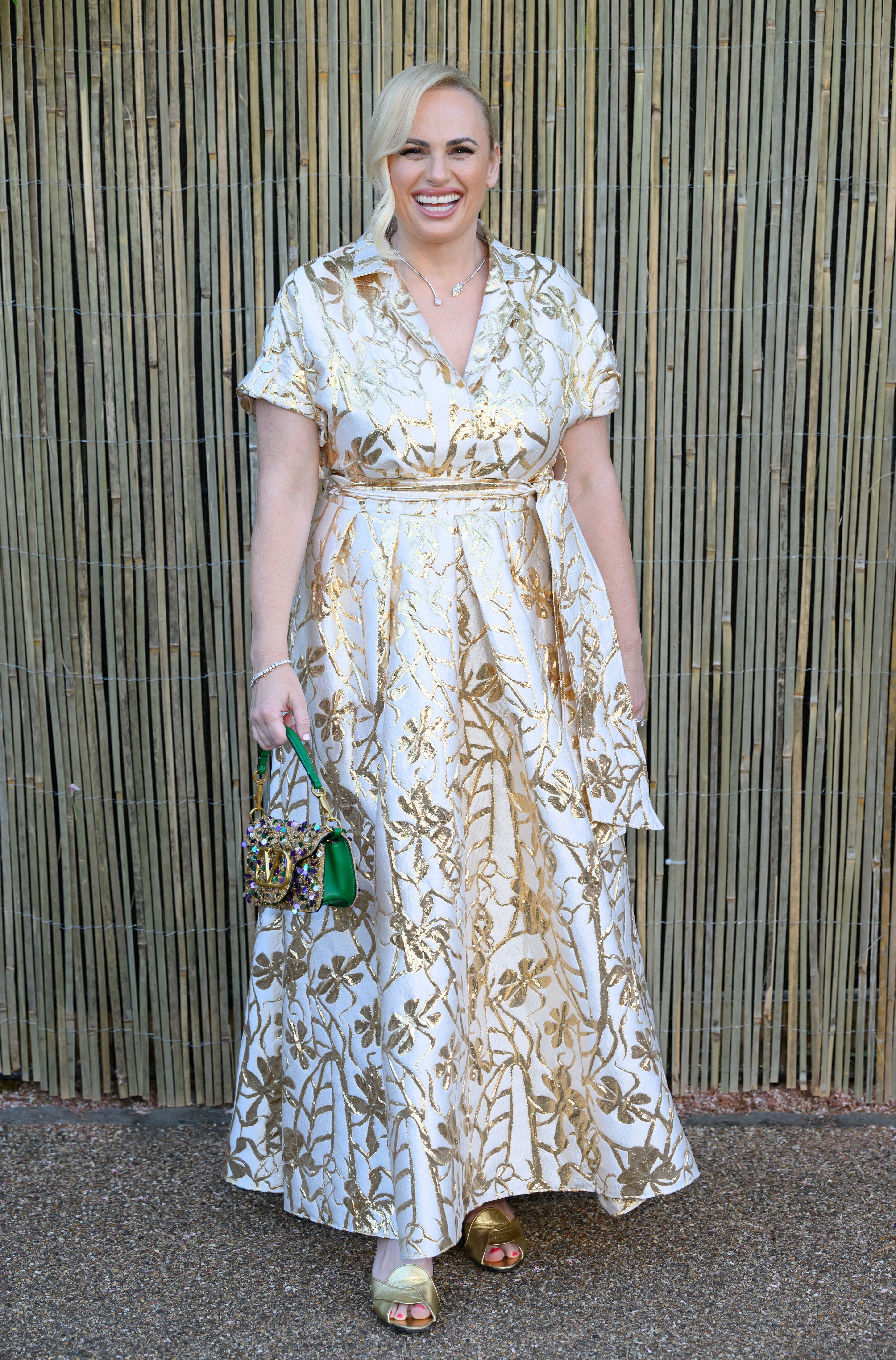 Rebel Wilson smiling, wearing a floral-patterned maxi dress, holding a small ornate bag in front of a bamboo backdrop