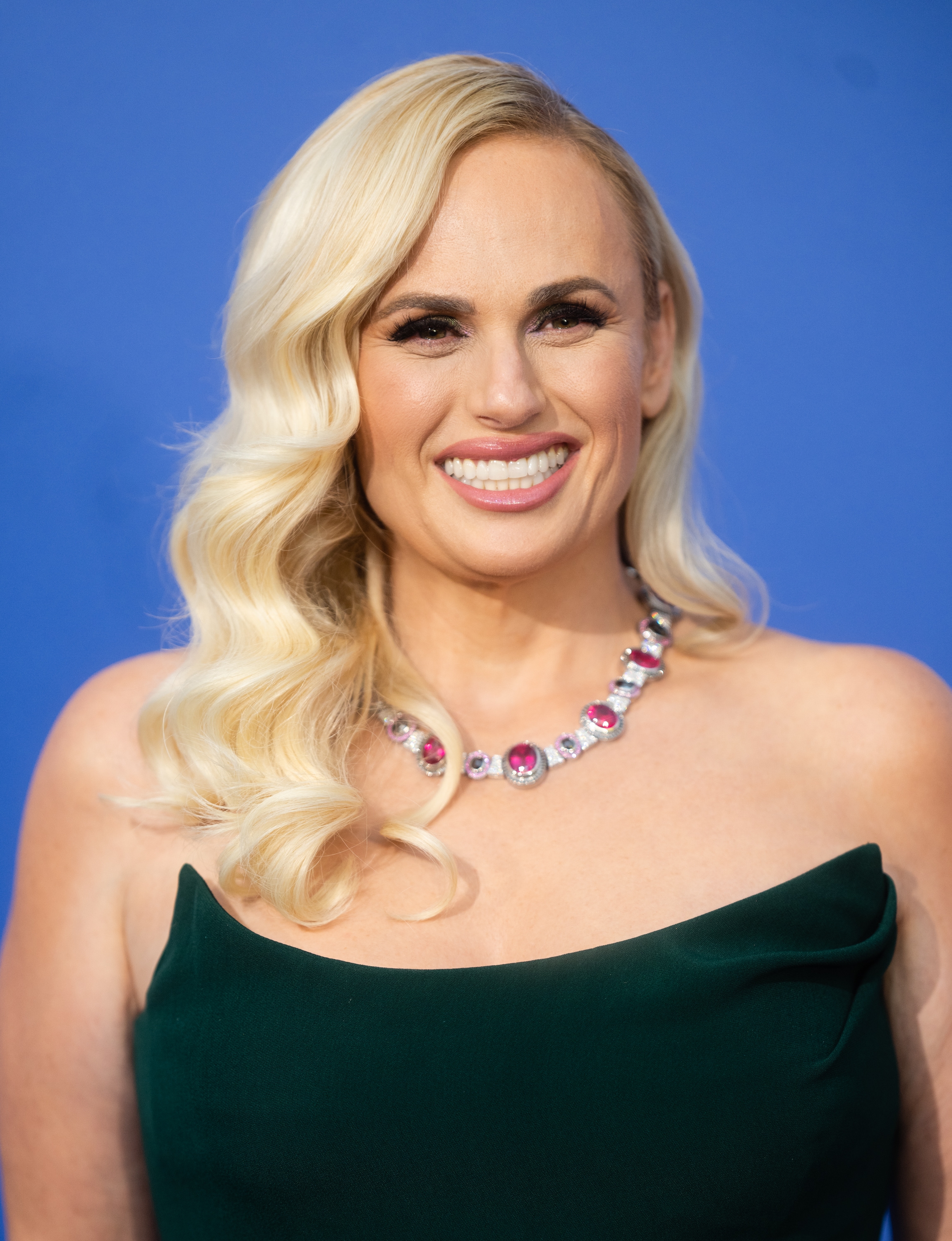 Rebel Wilson in strapless gown with wavy hair and statement necklace, smiling against a plain backdrop