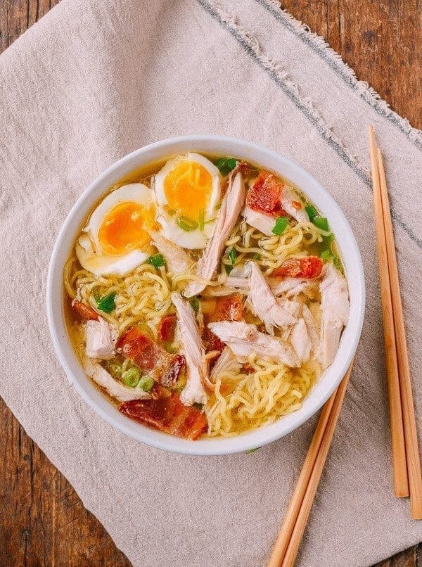 Bowl of ramen with sliced egg, chicken, bacon, and green onions on a cloth with chopsticks beside it