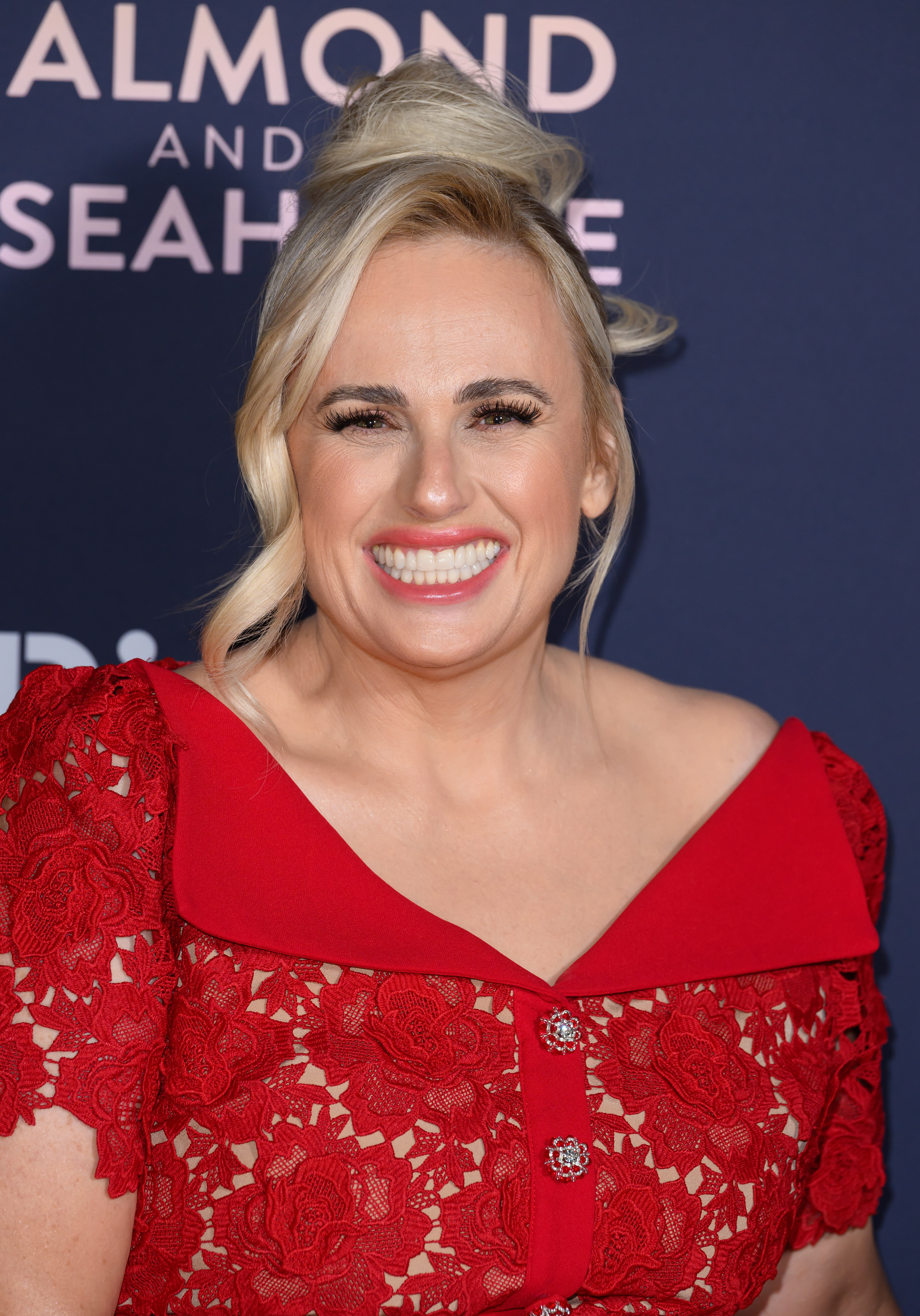 Rebel Wilson smiling on a red carpet, wearing a red lace dress with a boat neckline and decorative buttons