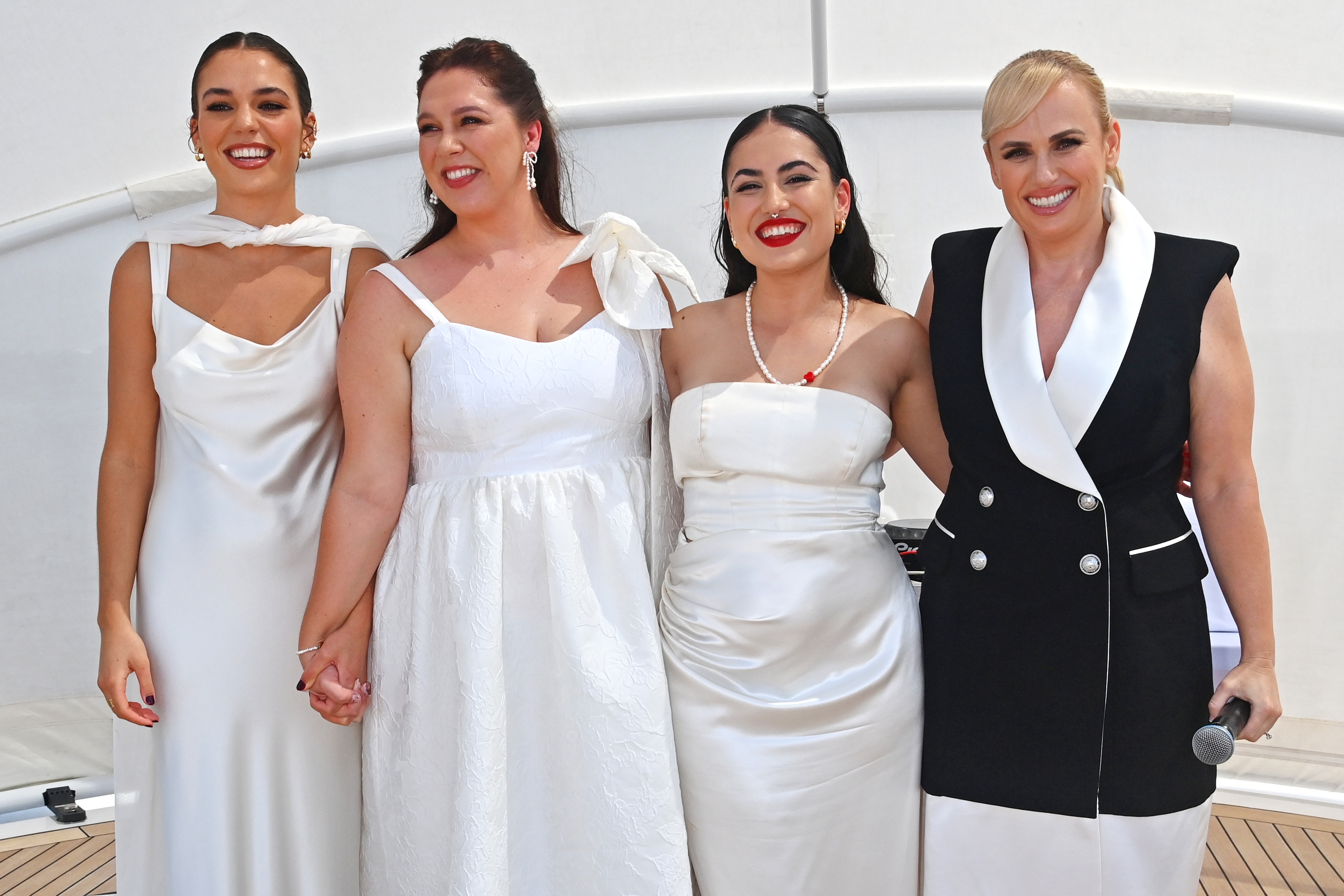 Four people pose together, smiling and holding hands in formal dresses and suits, with elegant accessories, at an outdoor event