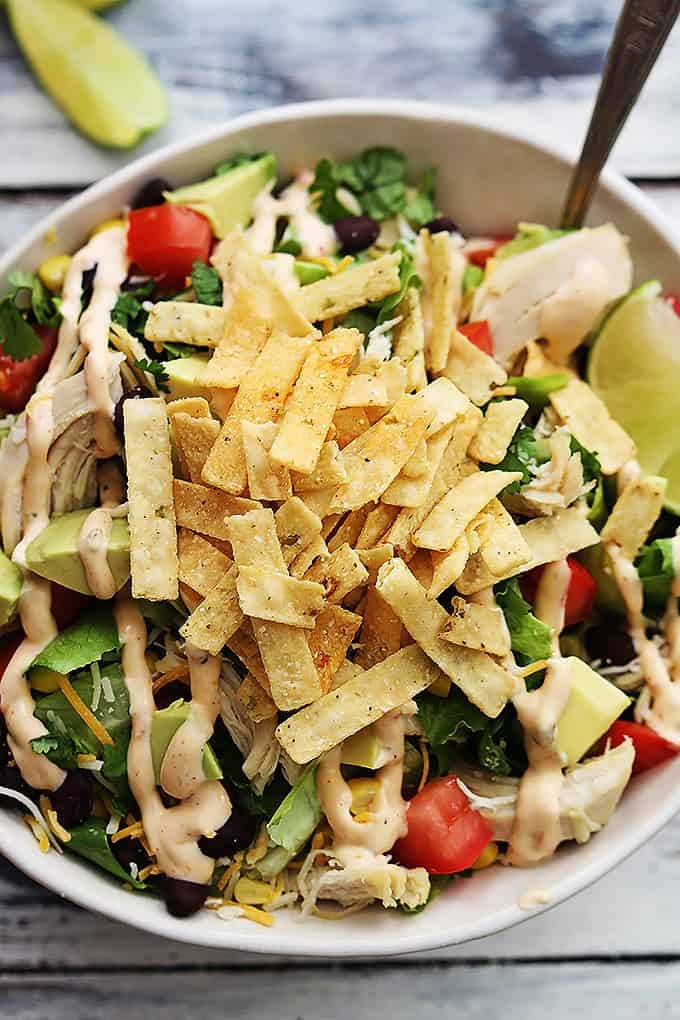 A bowl of salad topped with crispy tortilla strips, avocado, black beans, corn, tomatoes, lettuce, and drizzled with a creamy dressing