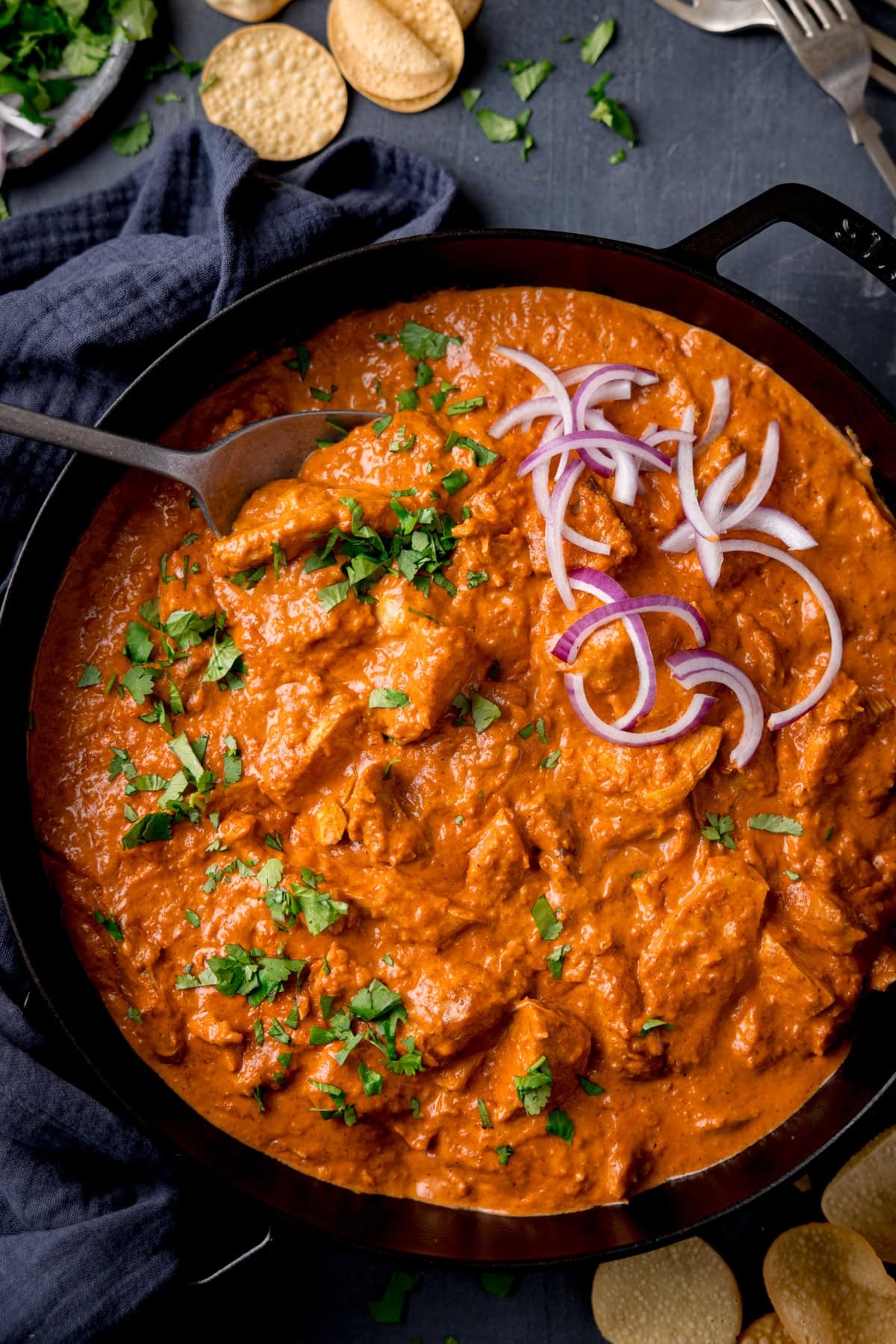 A bowl of creamy curry garnished with sliced onions and chopped herbs sits on a dark cloth with crackers nearby