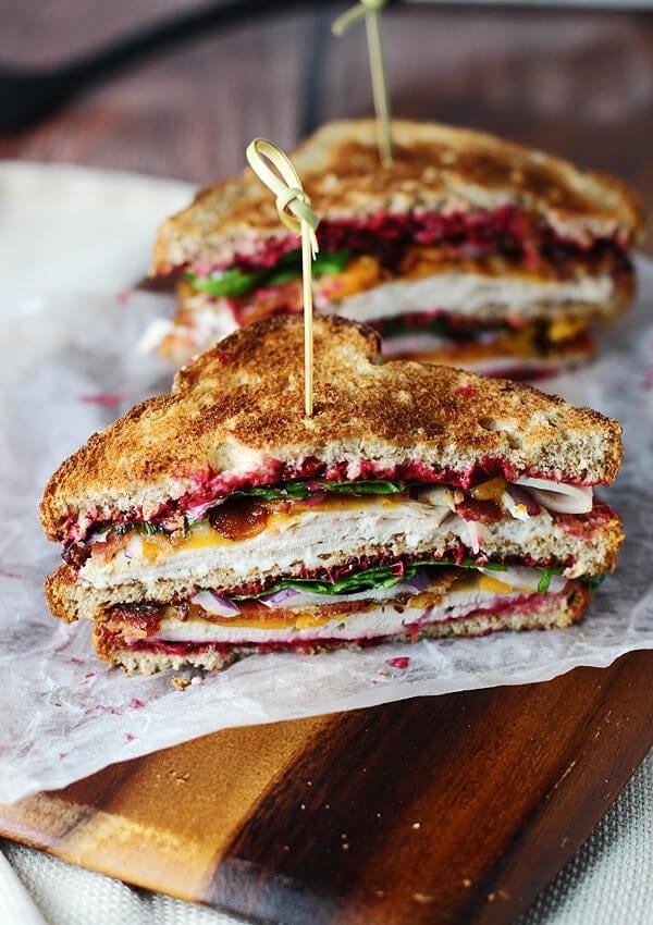 A club sandwich with layers of turkey, bacon, lettuce, and cranberry sauce, held together with a skewer, on a wooden board