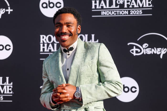 Donald Glover in a textured suit smiles on a red carpet at the Rock &amp;amp; Roll Hall of Fame Induction 2025 event. Logos visible in the background